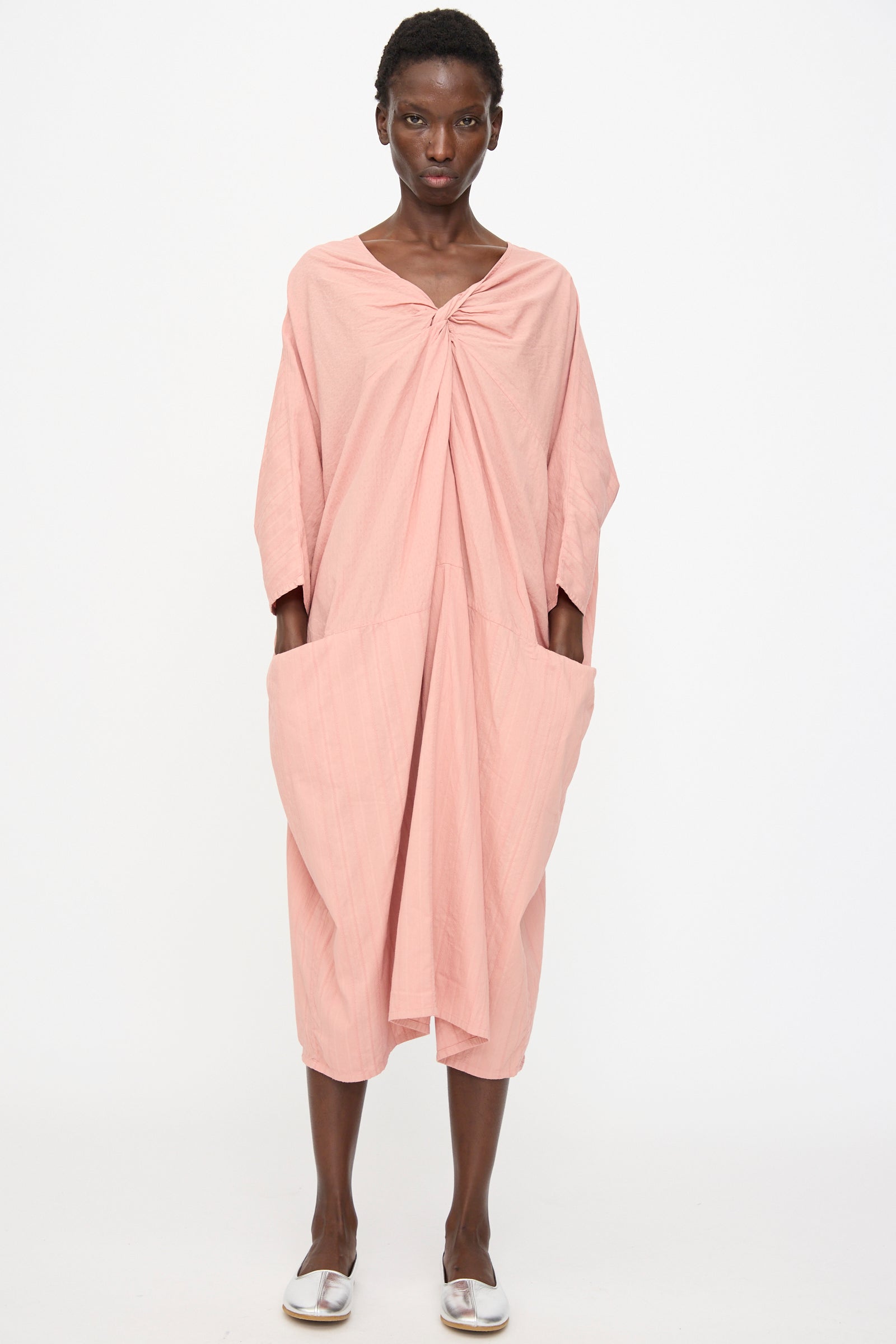 A person faces forward in Cosmic Wonder's Stripe Combination Twisted Dress in Rose Crystal—a relaxed fit with large pockets—paired with flat white shoes, all set against a plain white background.