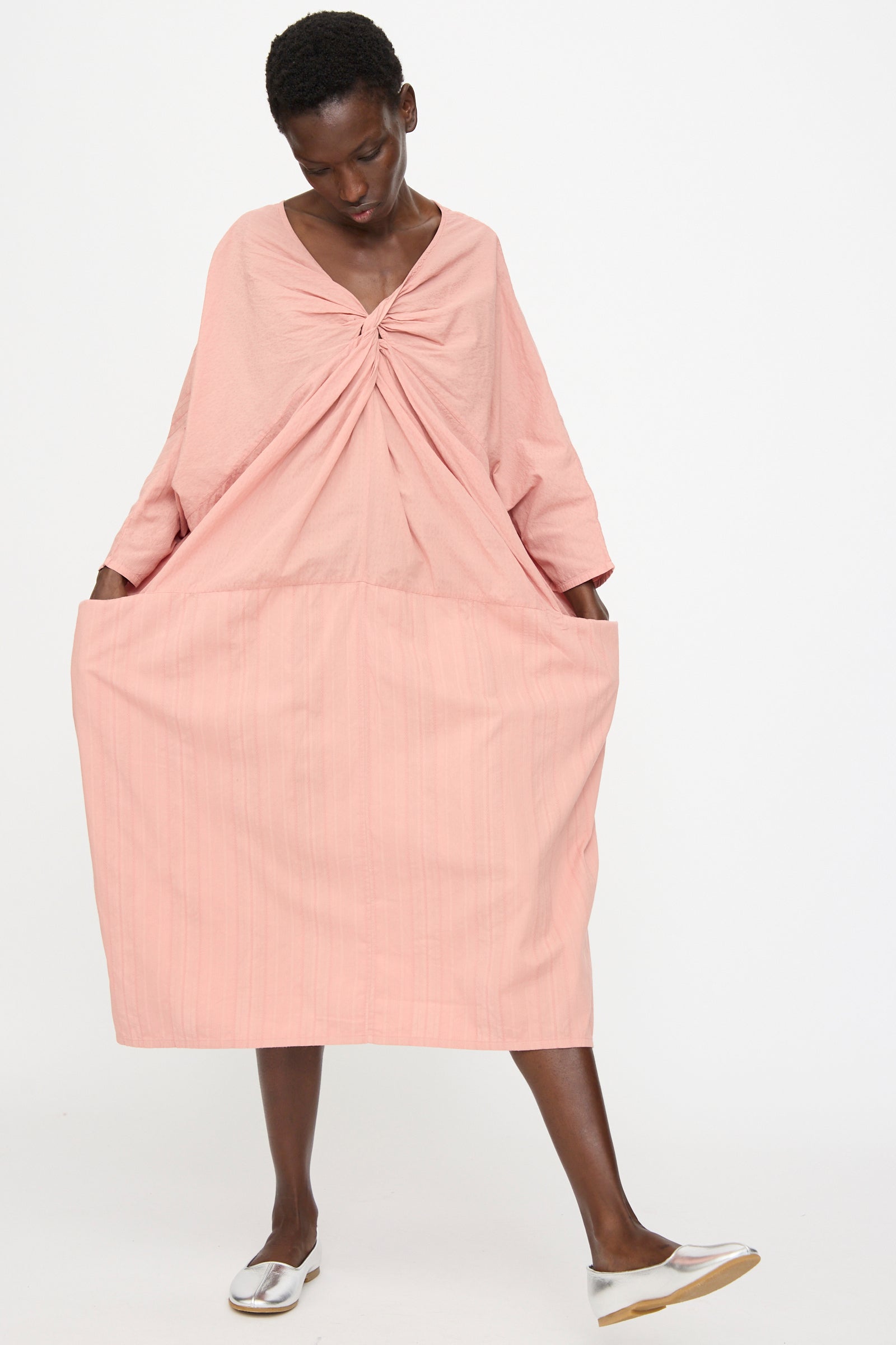 A person stands against a white background wearing the Cosmic Wonder Stripe Combination Twisted Dress in Rose Crystal—a relaxed-fit, loose pink dress with gathered front detail, large pockets, and silver flat shoes.