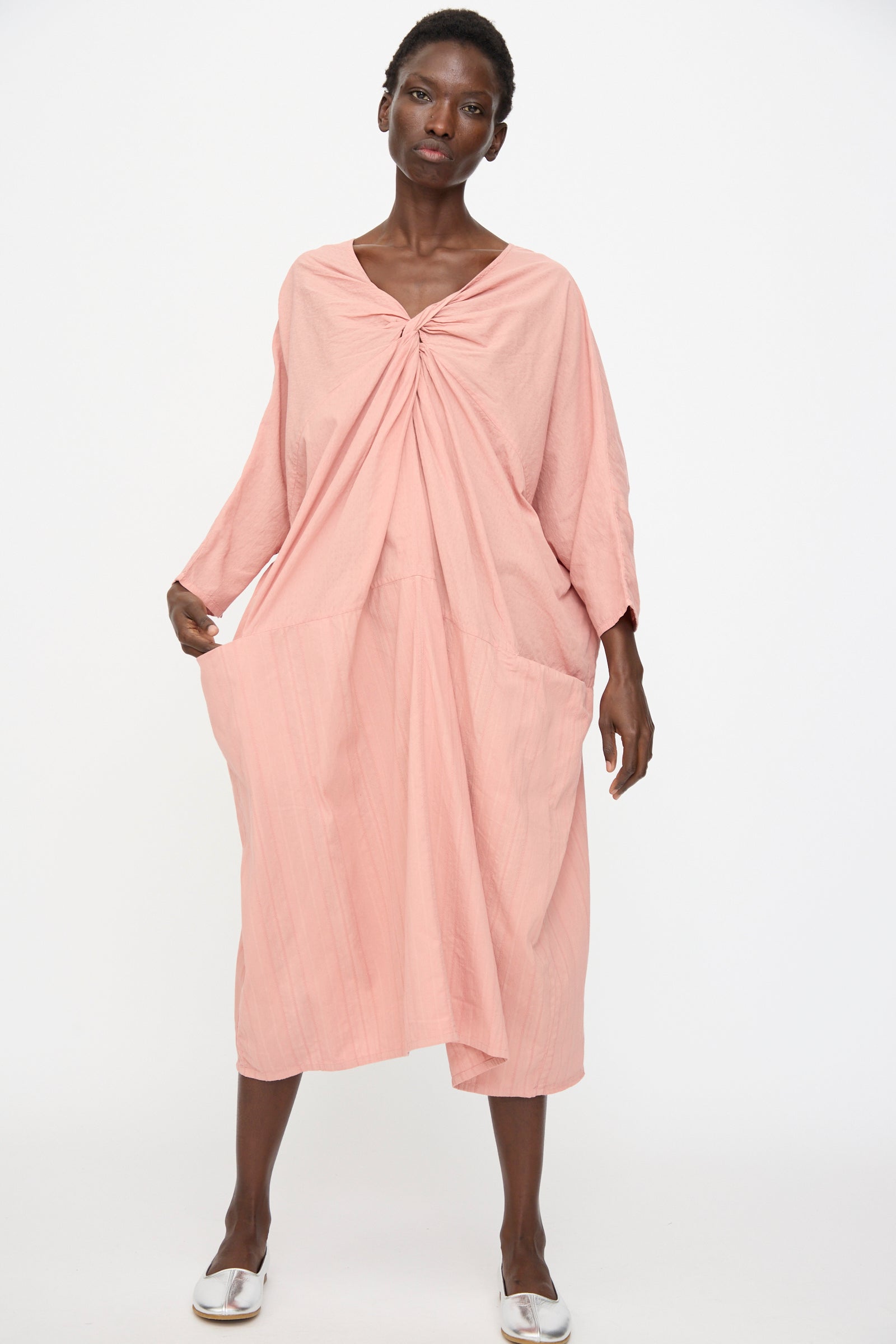 A person stands against a plain background wearing the Cosmic Wonder Stripe Combination Twisted Dress in Rose Crystal, featuring a relaxed fit and twisted knot detail at the neckline, paired with white shoes for subtle elegance.