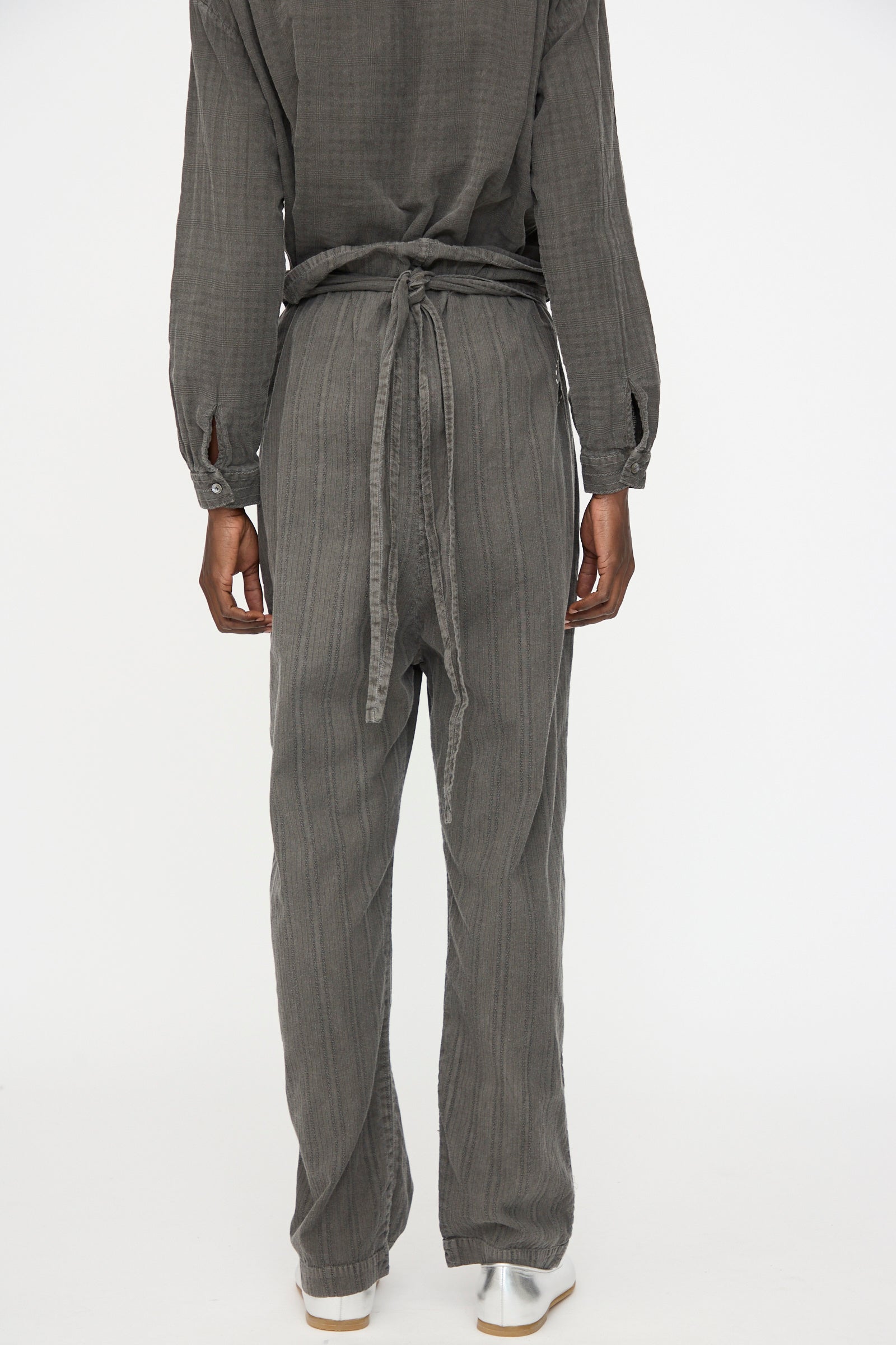 Person standing facing away, wearing Cosmic Wonder's Stripe Combination Wrapped Pant in Dark Sumi with a long-sleeved, gray textured top and white shoes, against a plain white background.