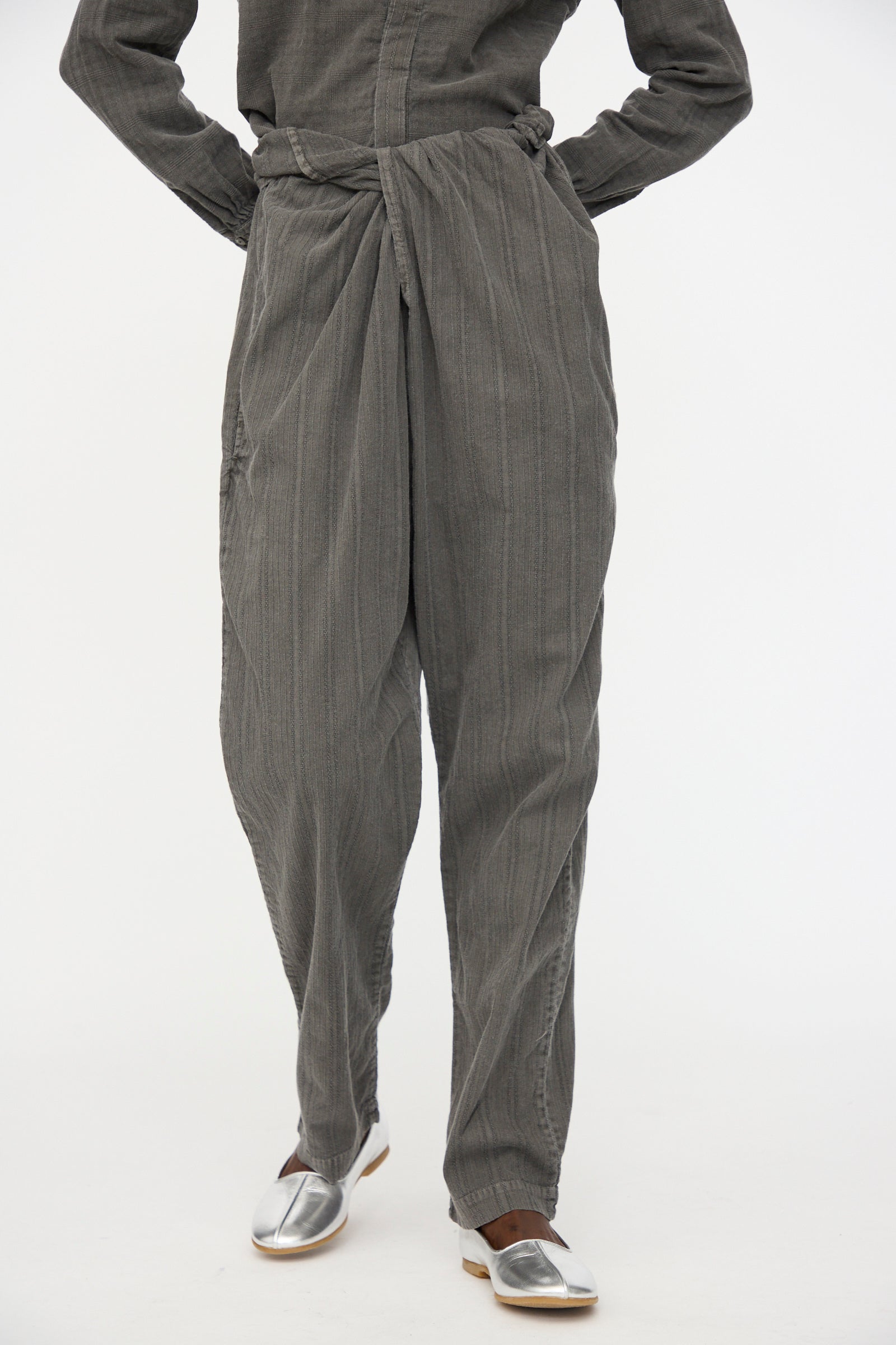 A person wears the Cosmic Wonder Stripe Combination Wrapped Pant in Dark Sumi—loose, textured gray organic cotton with a wrap waist—paired with silver slip-on shoes, standing against a plain white background.