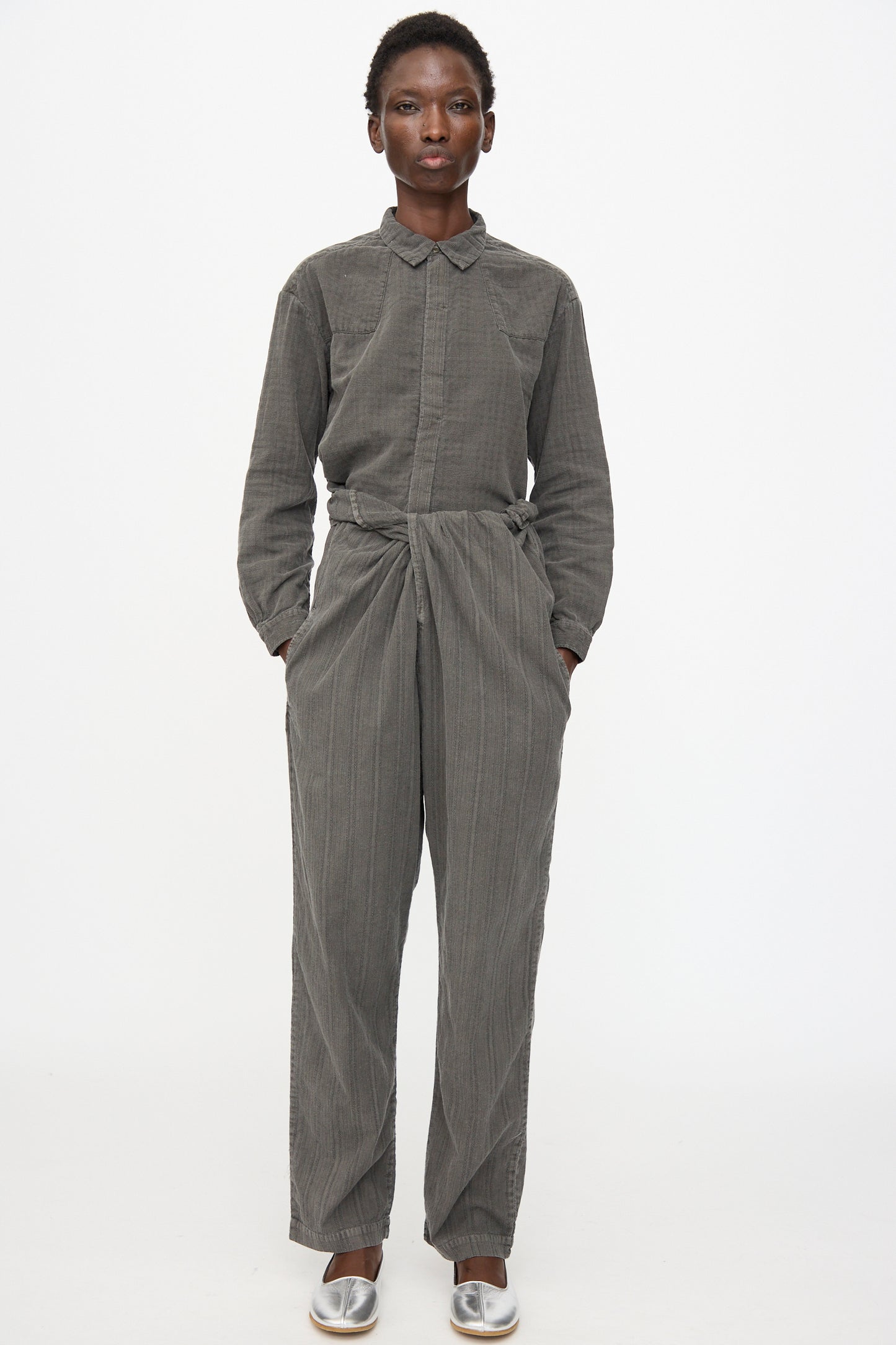 A person stands facing forward against a white background, wearing Cosmic Wonder's Stripe Combination Wrapped Pant in Dark Sumi with a grey long-sleeve collared top, silver shoes, and hands in pockets.