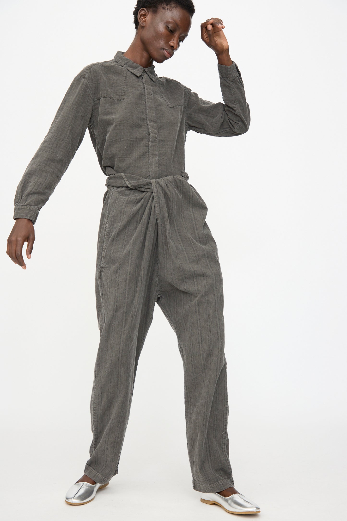 A person wears the Cosmic Wonder Stripe Combination Wrapped Pant in Dark Sumi, styled with a long-sleeve, button-up gray jumpsuit and silver shoes, standing against a plain white background.