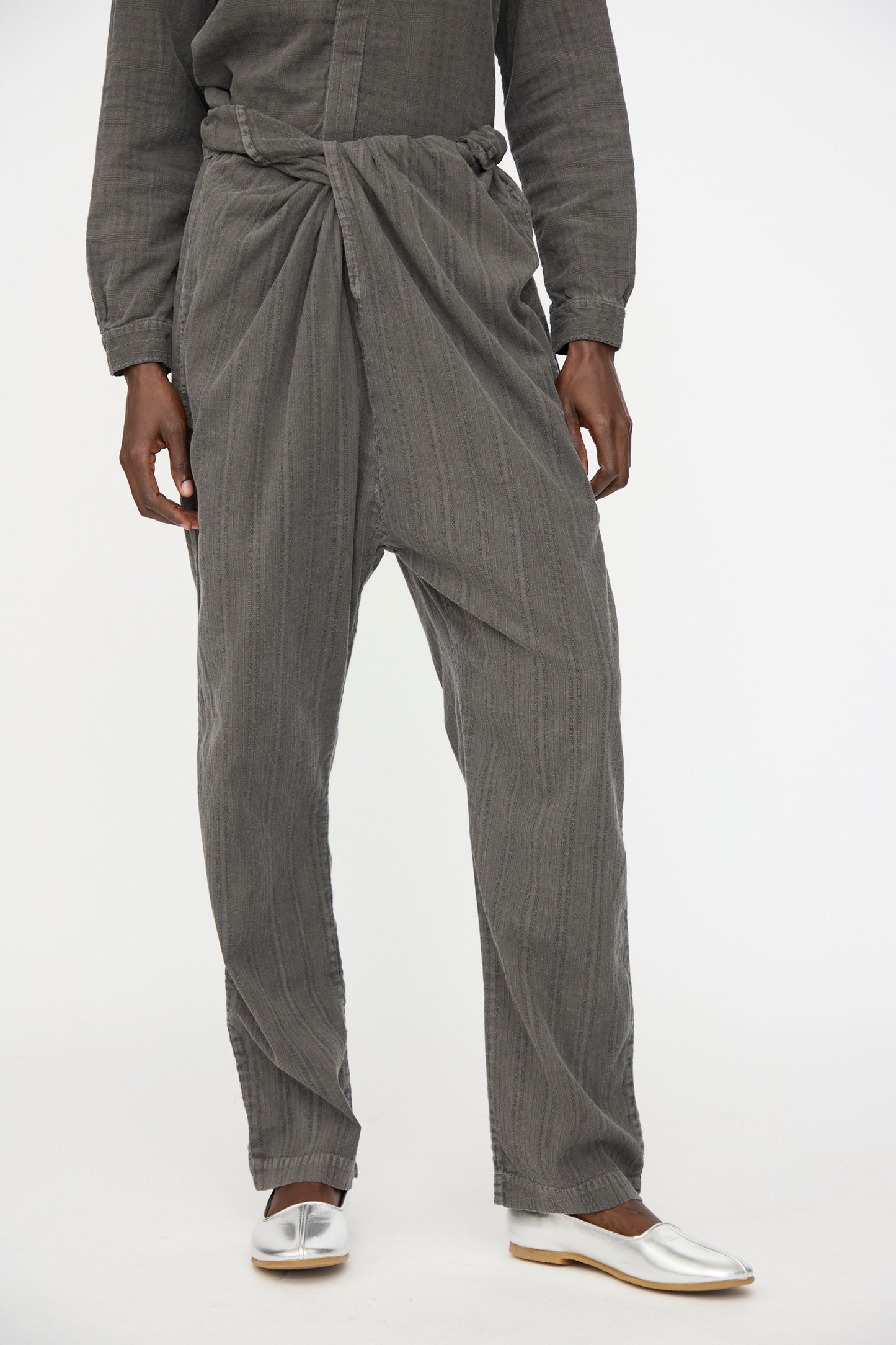 A person wears Cosmic Wonder’s Stripe Combination Wrapped Pant in Dark Sumi, styled with white shoes, standing against a plain white background.