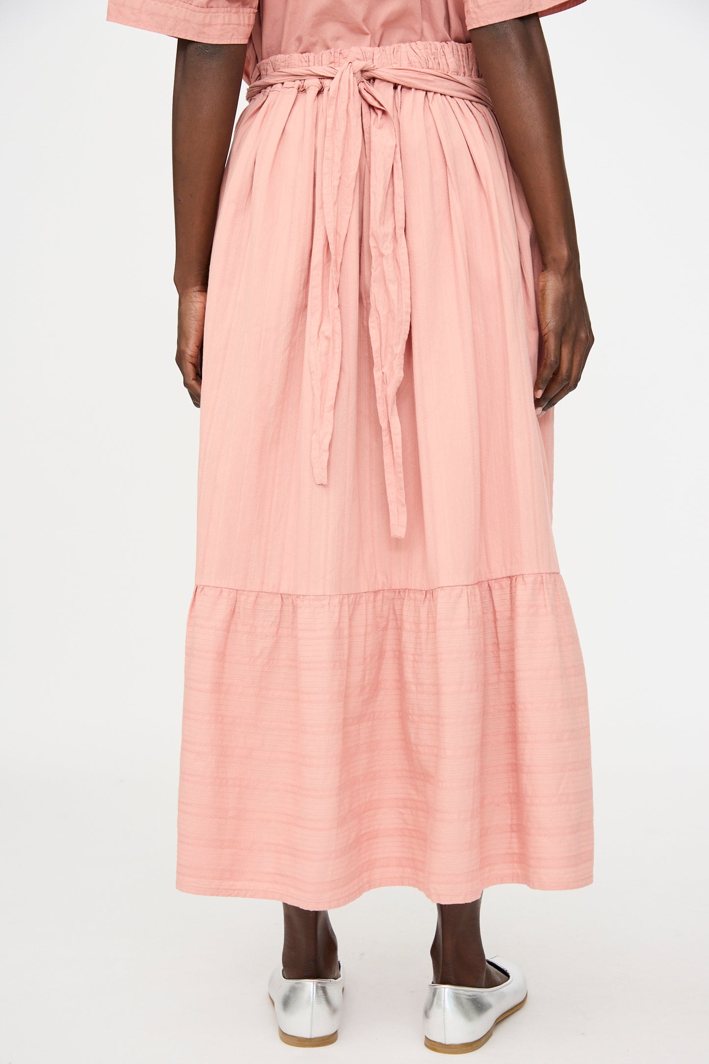 A person stands facing away, wearing the Cosmic Wonder Stripe Combination Wrapped Skirt in Rose Crystal paired with silver flat shoes.