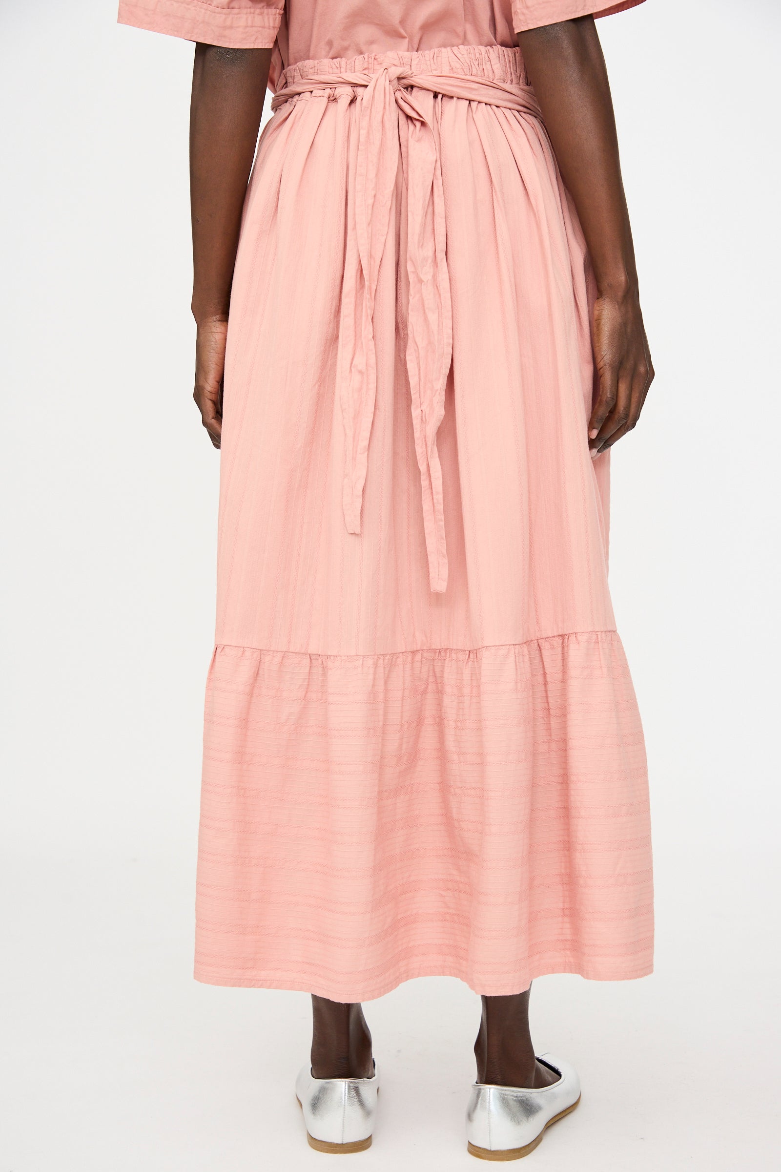 A person stands facing away, wearing the Cosmic Wonder Stripe Combination Wrapped Skirt in Rose Crystal paired with silver flat shoes.