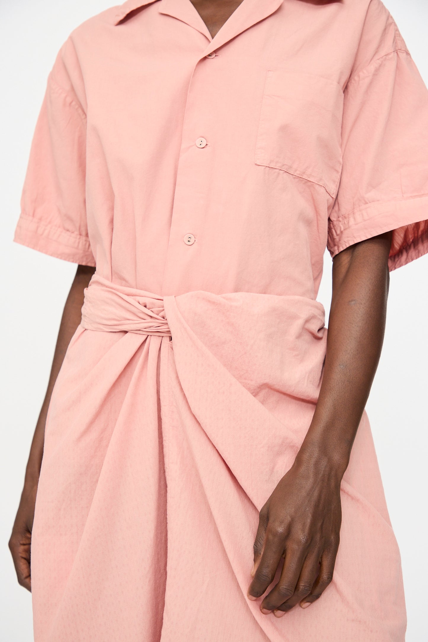 A person wears a light pink short-sleeve button-up dress with waist knot and pocket, paired with Cosmic Wonder's Stripe Combination Wrapped Skirt in Rose Crystal.
