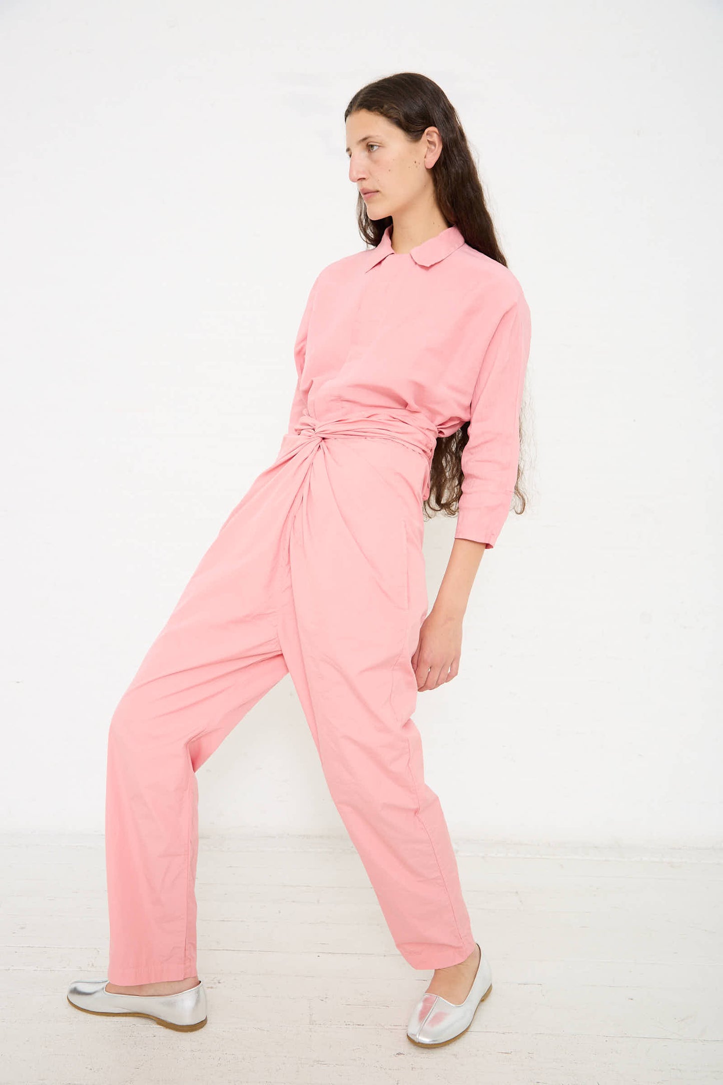 A woman with long hair wearing a pink button-up shirt and high-waisted Cosmic Wonder Suvin Cotton Broadcloth Pant in Peach Jade with an adjustable tie waist poses against a plain white background.