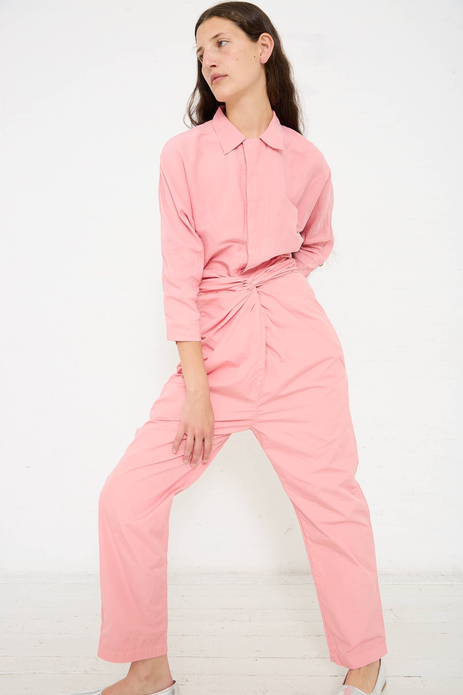 Person in a pink, long-sleeved shirt and matching Cosmic Wonder Suvin Cotton Broadcloth Pant in Peach Jade with an adjustable tie waist stands against a white background, posing with one leg bent and hands relaxed by their sides.