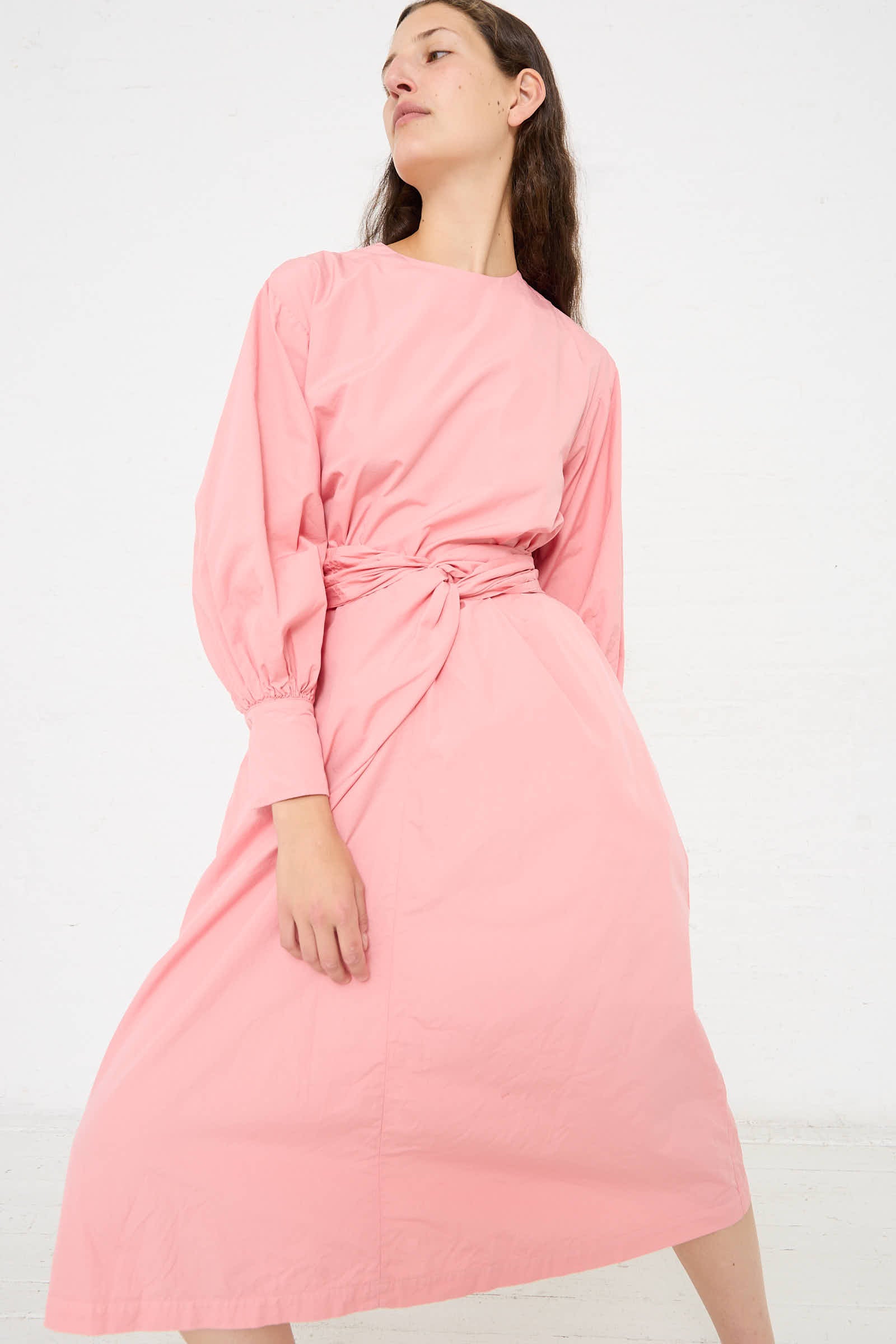 Person standing against a white background, wearing a loose-fitting, long-sleeved, Suvin Cotton Broadcloth Wrapped Dress in Peach Jade by Cosmic Wonder.