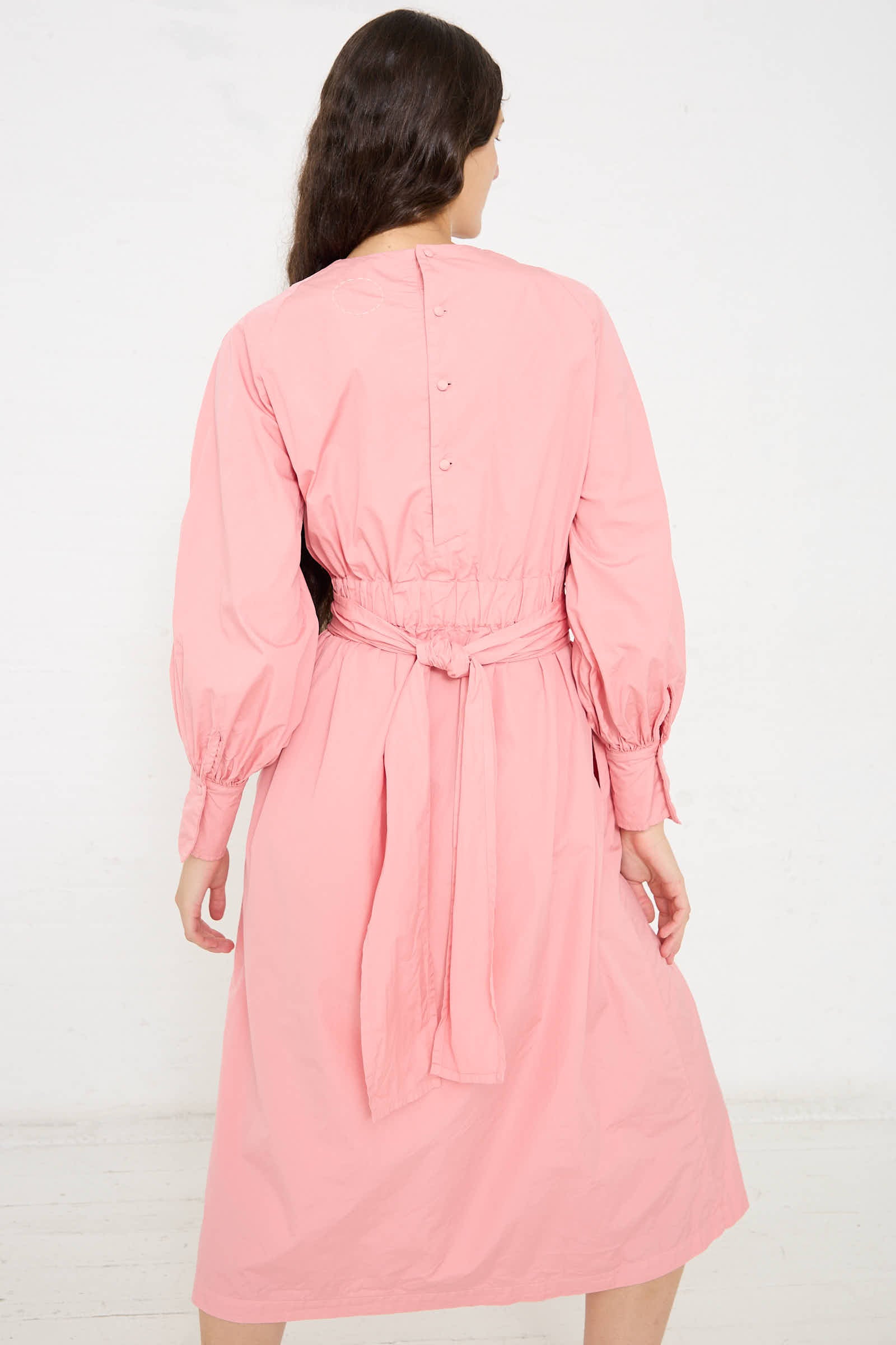 A person with long dark hair is standing and facing away, wearing a long-sleeved, belted Suvin Cotton Broadcloth Wrapped Dress in Peach Jade by Cosmic Wonder with buttons on the upper back.