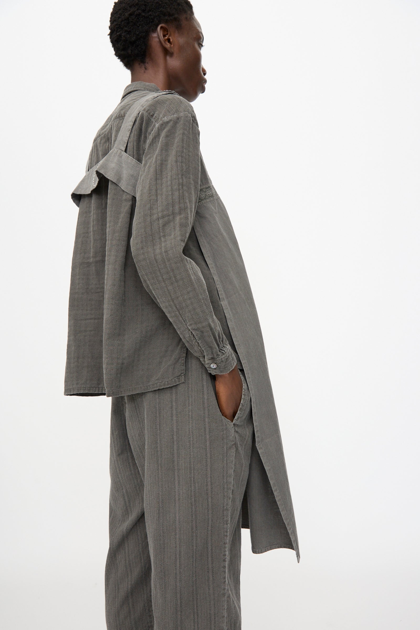 A person stands sideways against a plain white background, wearing Cosmic Wonder's Taskuki Apron in Dark Sumi—an antique-inspired, loose-fitting gray pinstripe set with fabric detail on the back and matching wide-leg trousers.