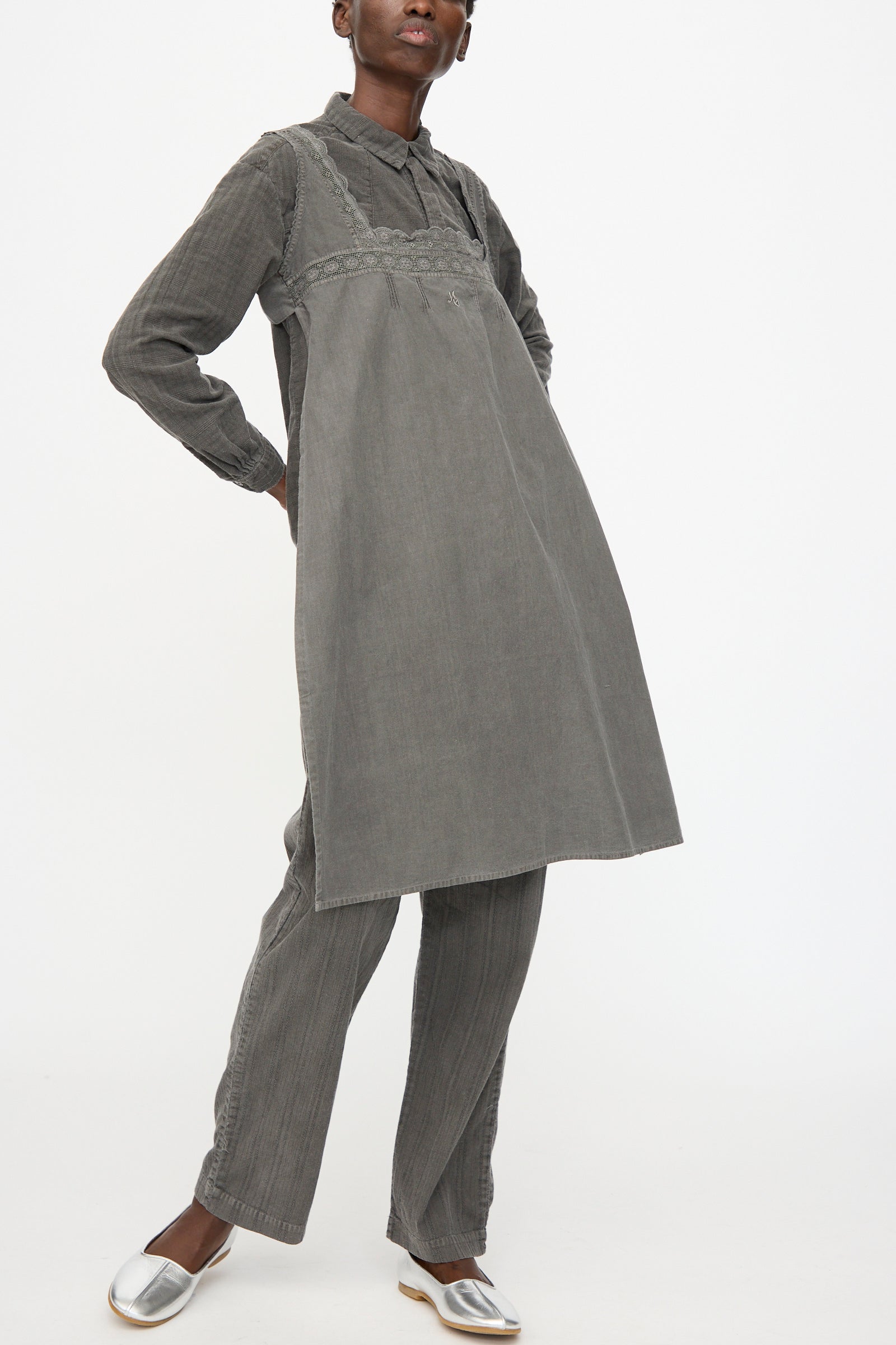A person wears the Cosmic Wonder Taskuki Apron in Dark Sumi, styled over a gray long-sleeved shirt, matching gray pants, and silver shoes, standing against a plain white background.