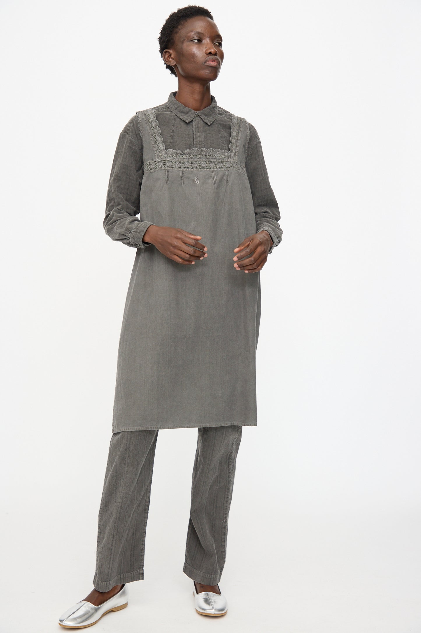 A person stands against a plain white background wearing Cosmic Wonder's Taskuki Apron in Dark Sumi, styled over grey layers and silver shoes.