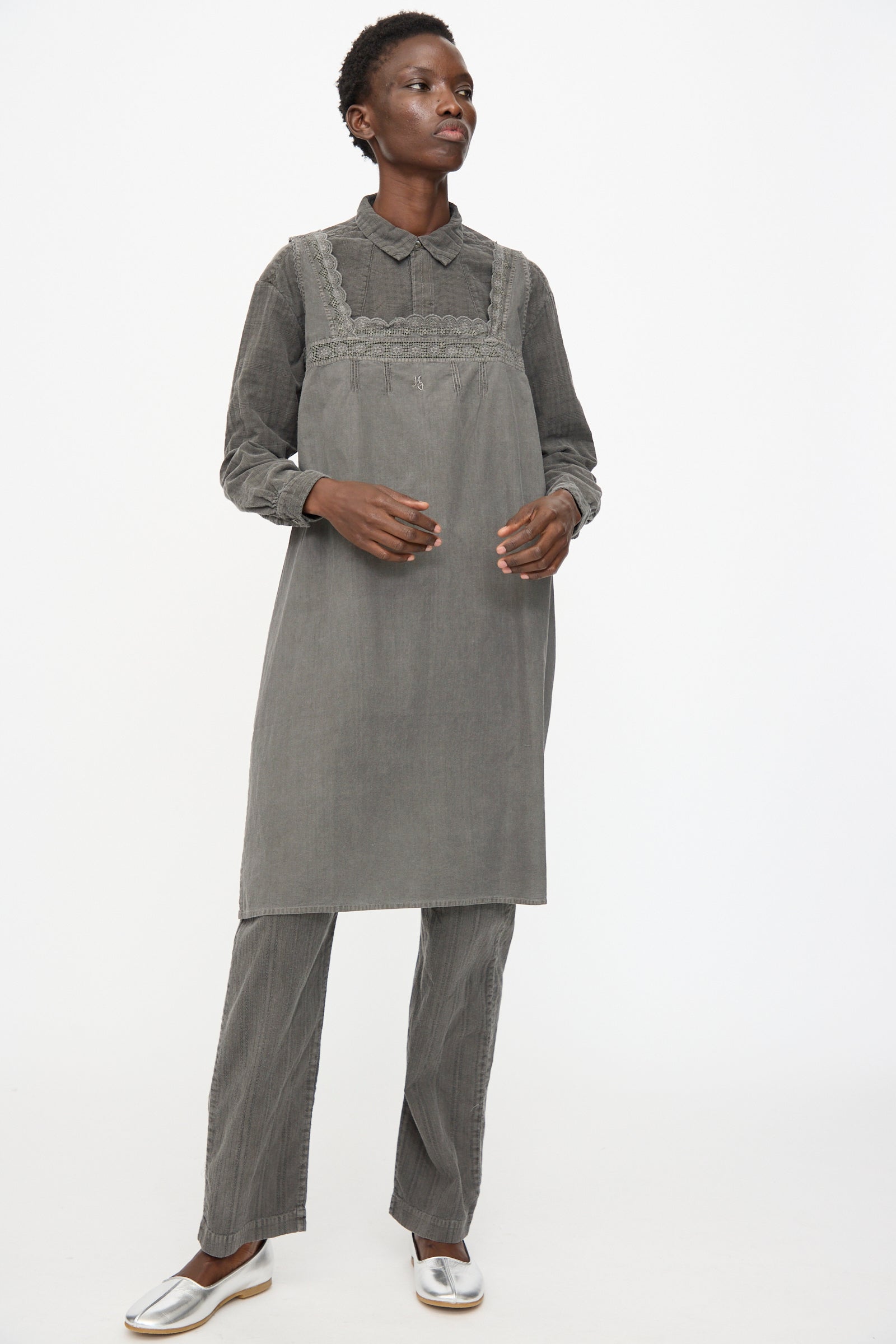 A person stands against a plain white background wearing Cosmic Wonder's Taskuki Apron in Dark Sumi, styled over grey layers and silver shoes.