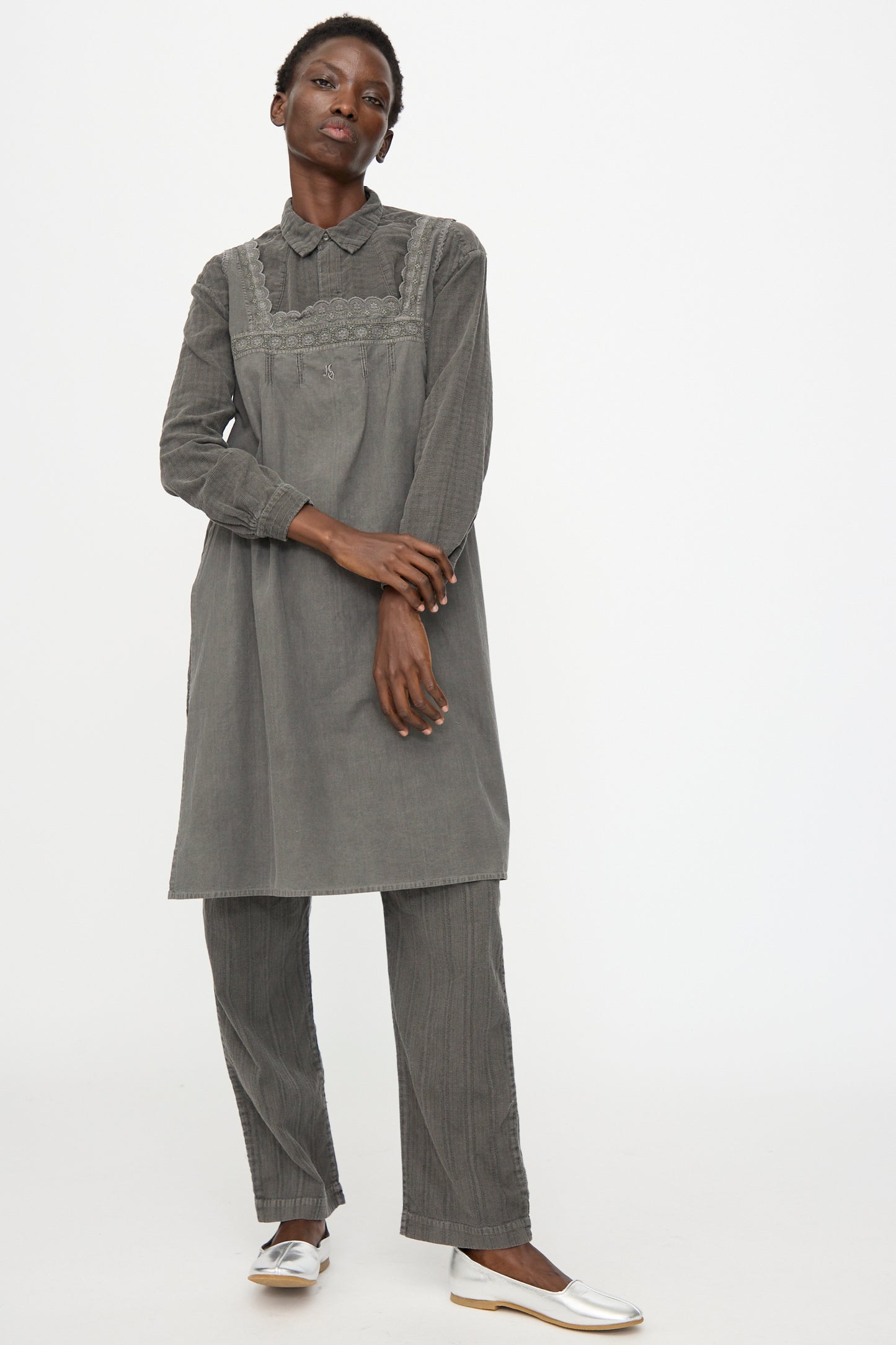 A person stands against a plain background wearing the Cosmic Wonder Taskuki Apron in Dark Sumi, a loose-fitting, long-sleeved gray tunic over matching pants and white flat shoes, inspired by antique Japanese undergarments.