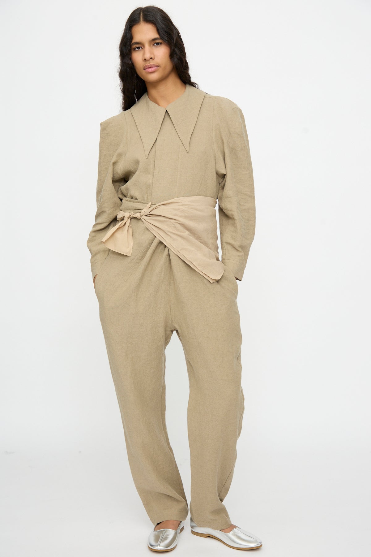 A model wears a tan, long-sleeved jumpsuit with a wide, pointed collar and a Cosmic Wonder The Farmer’s Star Belt in Amber Light tied at the waist.