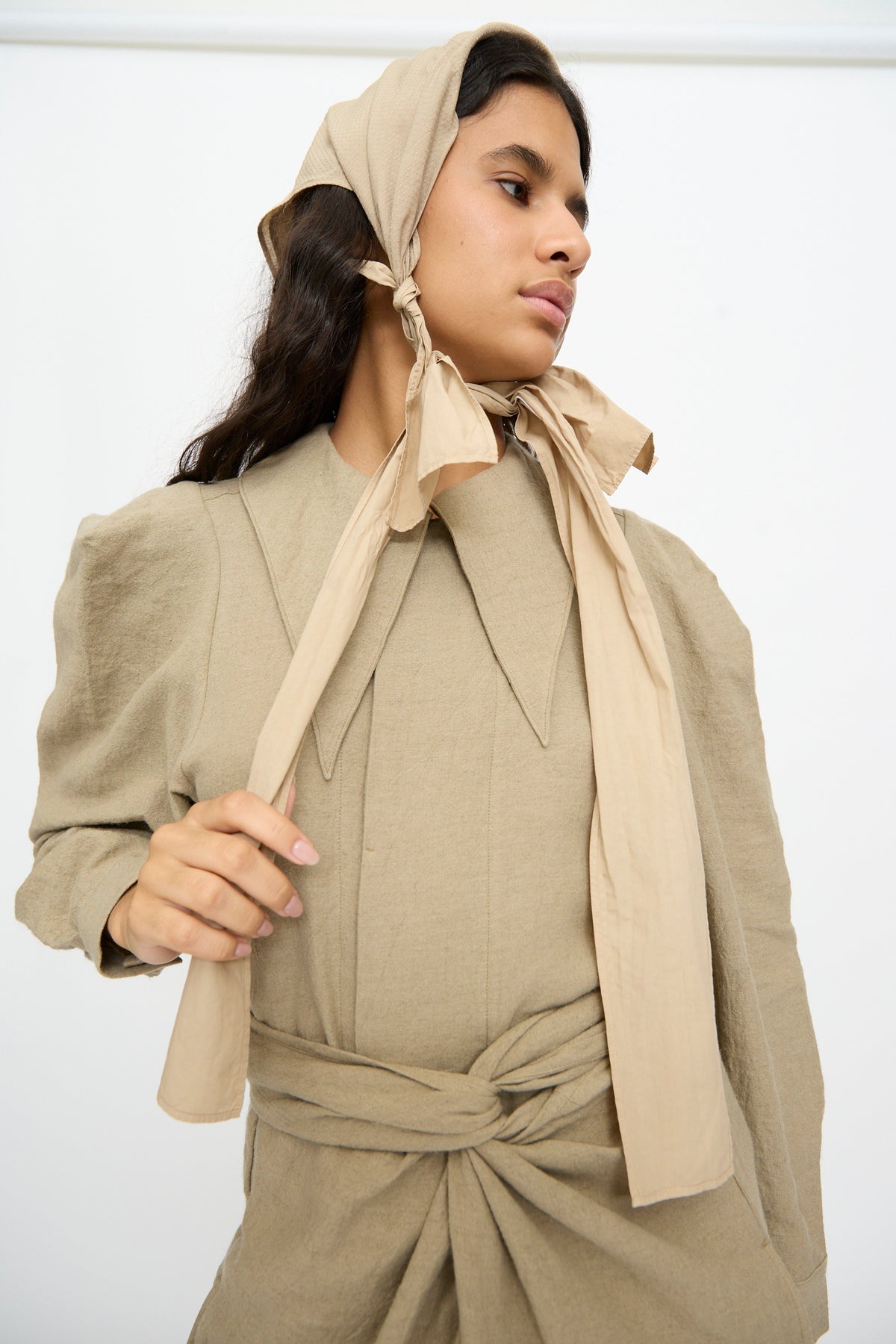 A person wears a tan headscarf tied under the chin and a matching long-sleeved dress with a large collar and The Farmer’s Star Belt in Amber Light.