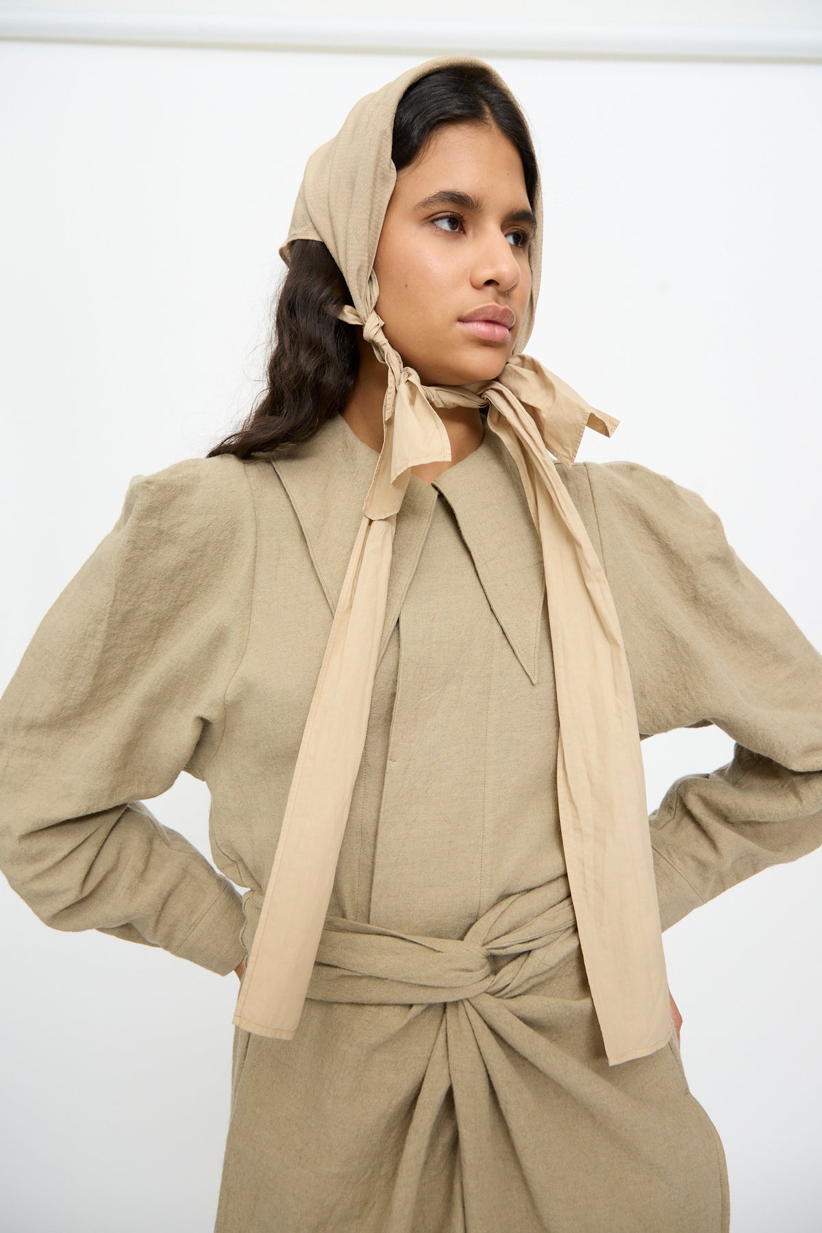 A person wears a beige headscarf tied under the chin and a matching long-sleeved dress with a wide collar and The Farmer’s Star Belt in Amber Light tied at the waist.
