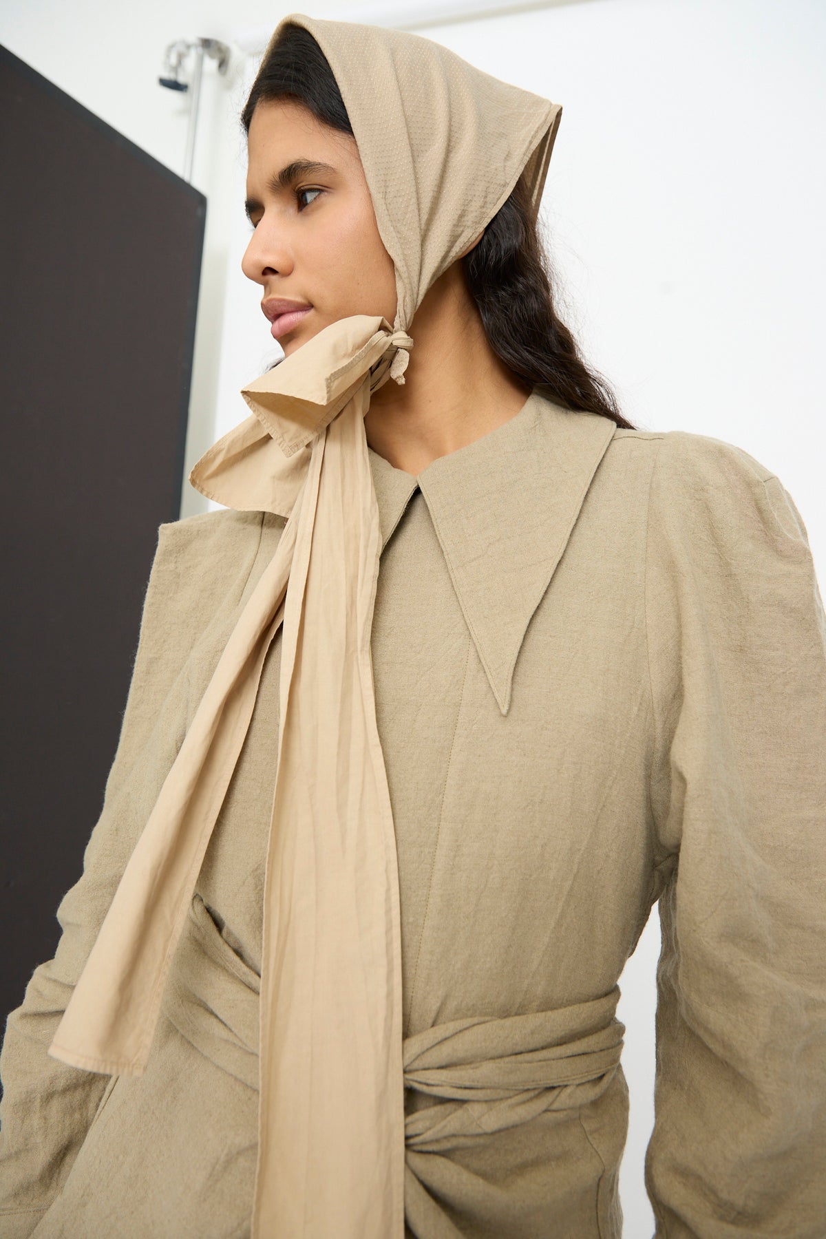 A person wearing a tan headscarf tied under the chin and a matching long-sleeved dress with a large collar and The Farmer’s Star Belt in Amber Light tied at the waist.