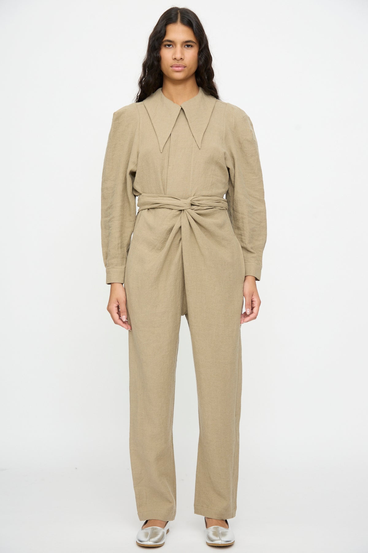 A person wearing a Cosmic Wonder Wrap Pants in beige, featuring a long-sleeved wool blend jumpsuit with a pointed collar and a knotted waist tie.