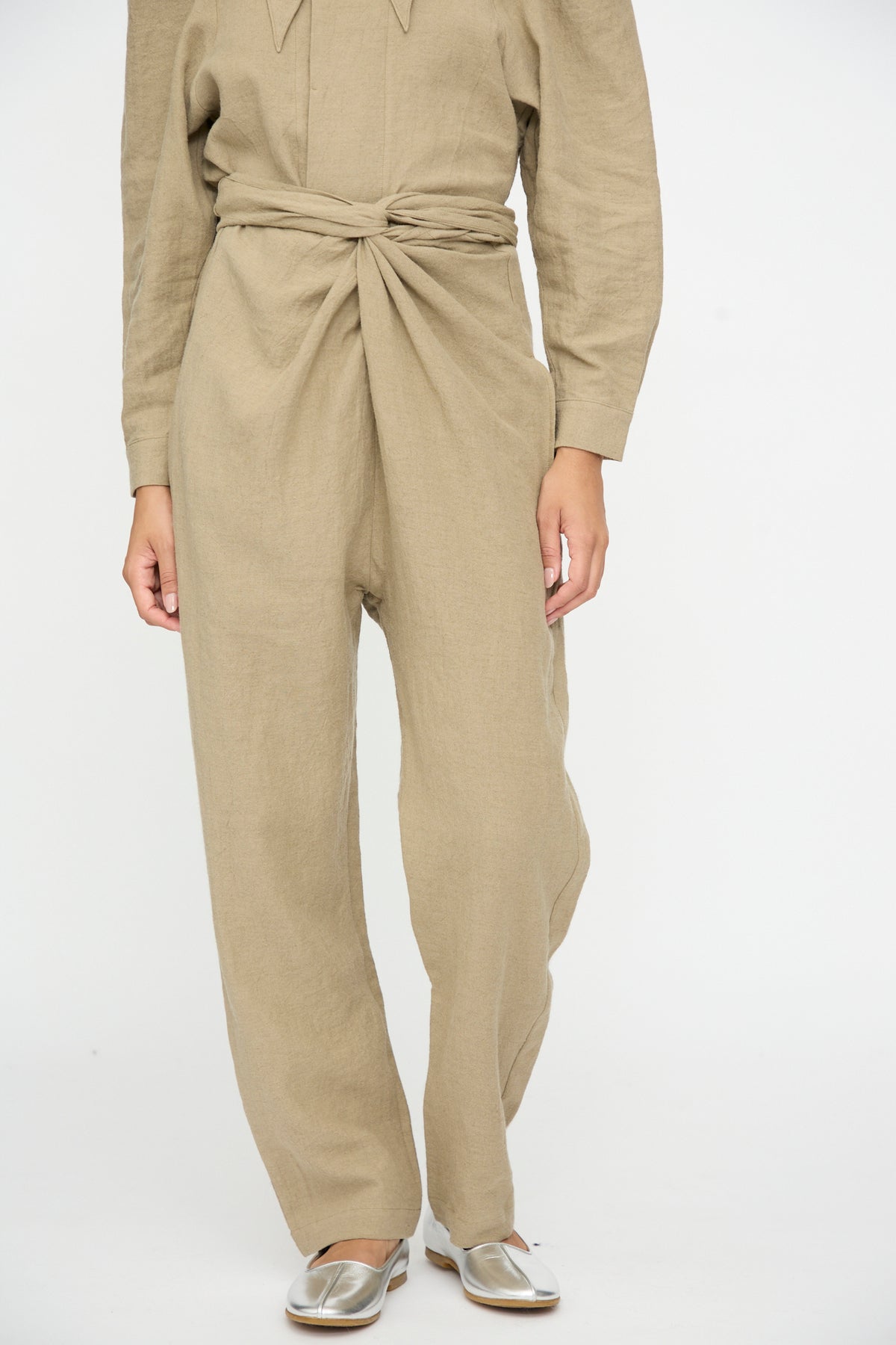 A person wearing Cosmic Wonder Wrap Pants in Beige tied at the waist and silver flat shoes.