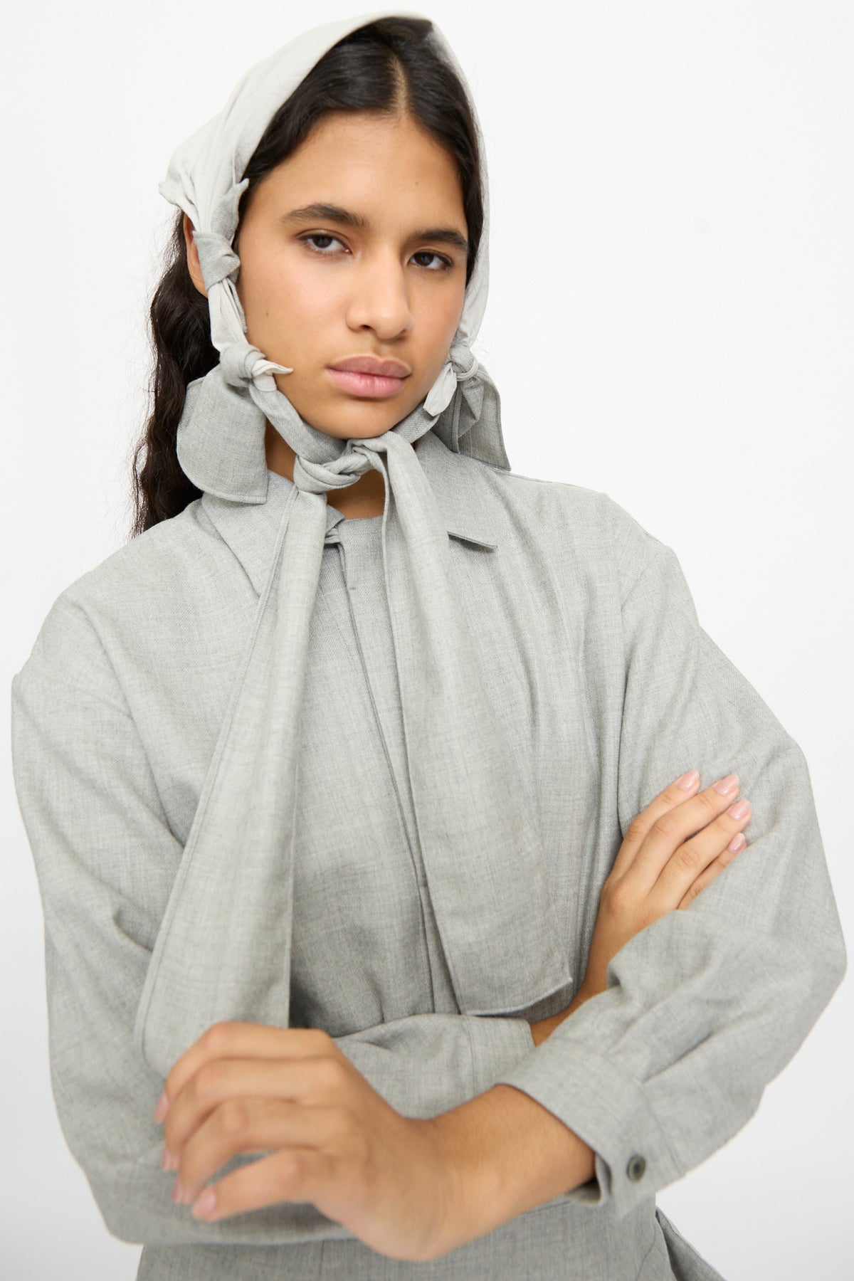 A person wearing a gray wool and cotton button-down shirt and matching headscarf tied under the chin, accessorized with the Nostalgic Painter’s Star Belt in Gray, shown with arms crossed.