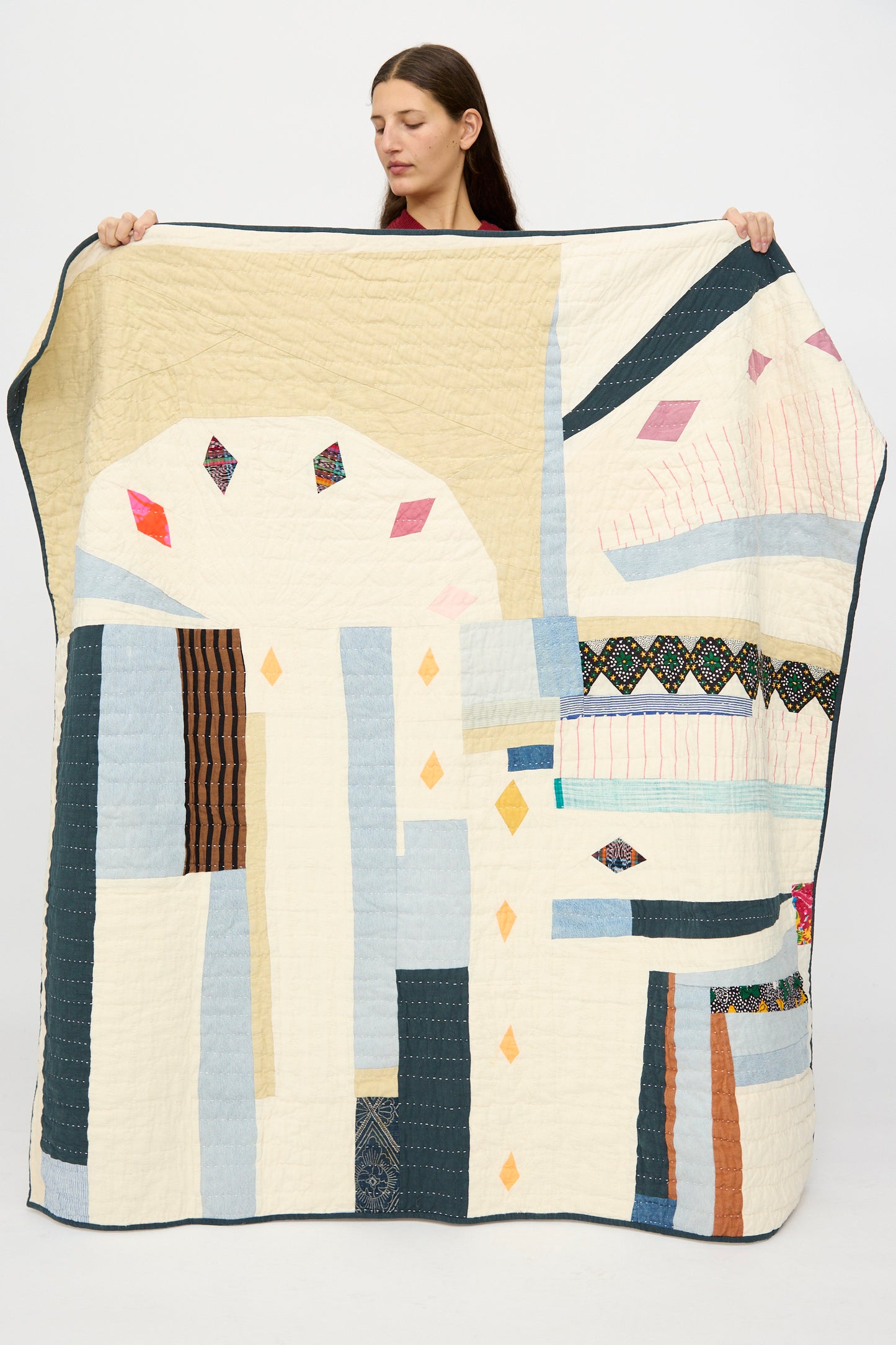 A woman stands against a plain background, holding up the Counterpane Large Patchwork Quilt in Cream Multi, handmade from found cloth and featuring abstract geometric patterns with various colors and textures.