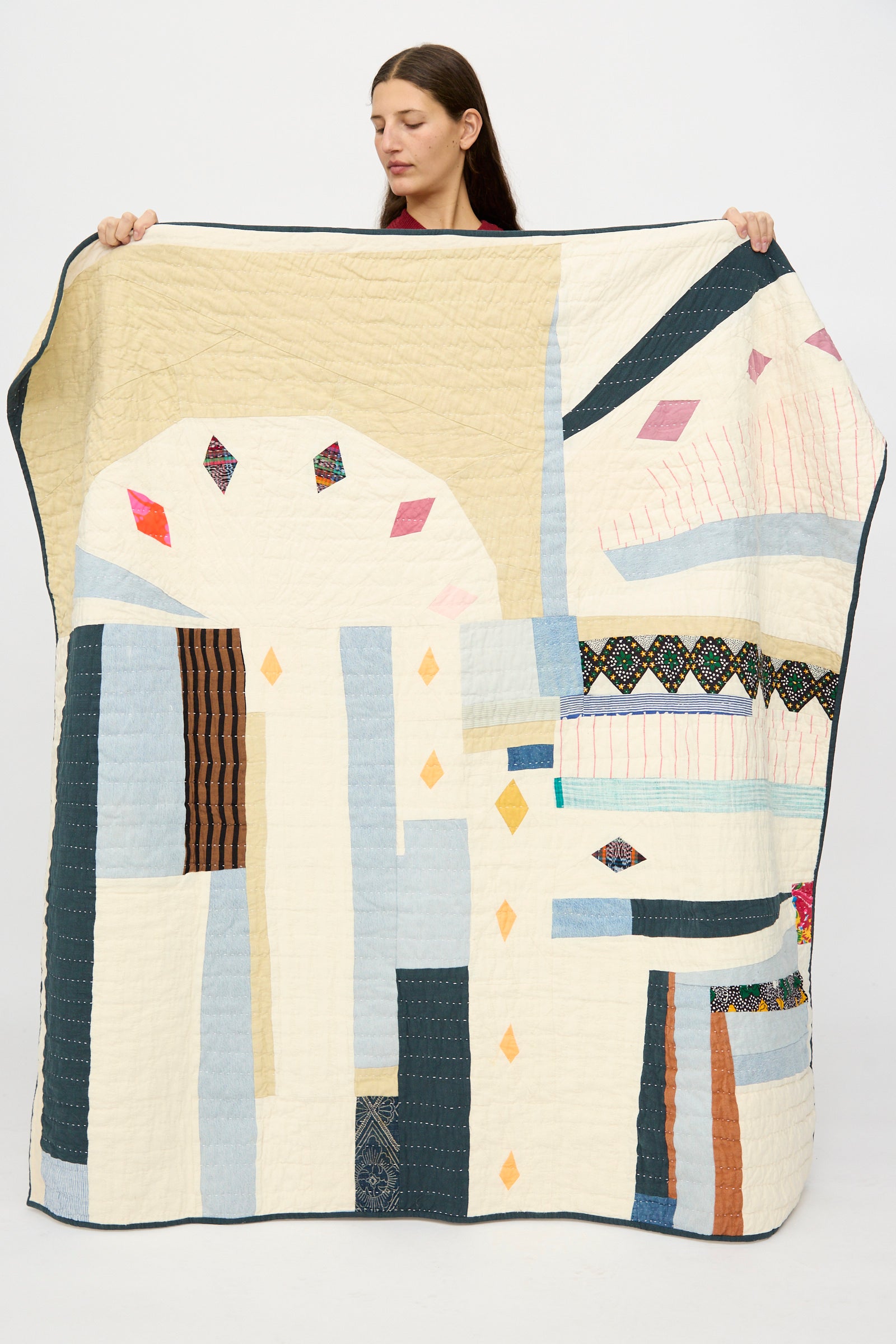A woman stands against a plain background, holding up the Counterpane Large Patchwork Quilt in Cream Multi, handmade from found cloth and featuring abstract geometric patterns with various colors and textures.