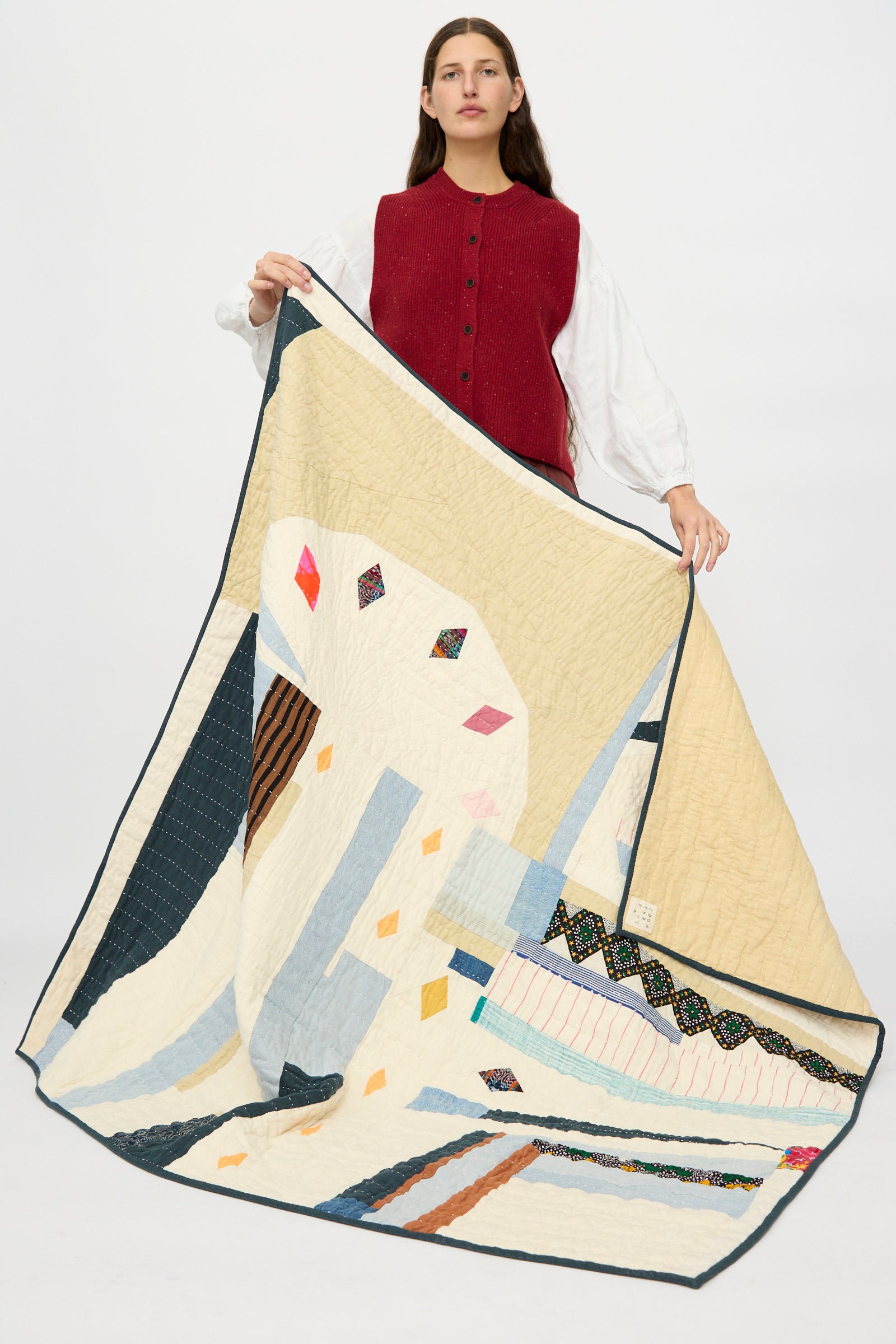 A woman in a red vest and white shirt holds the Counterpane Large Patchwork Quilt in Cream Multi, featuring colorful geometric shapes, against a plain background.