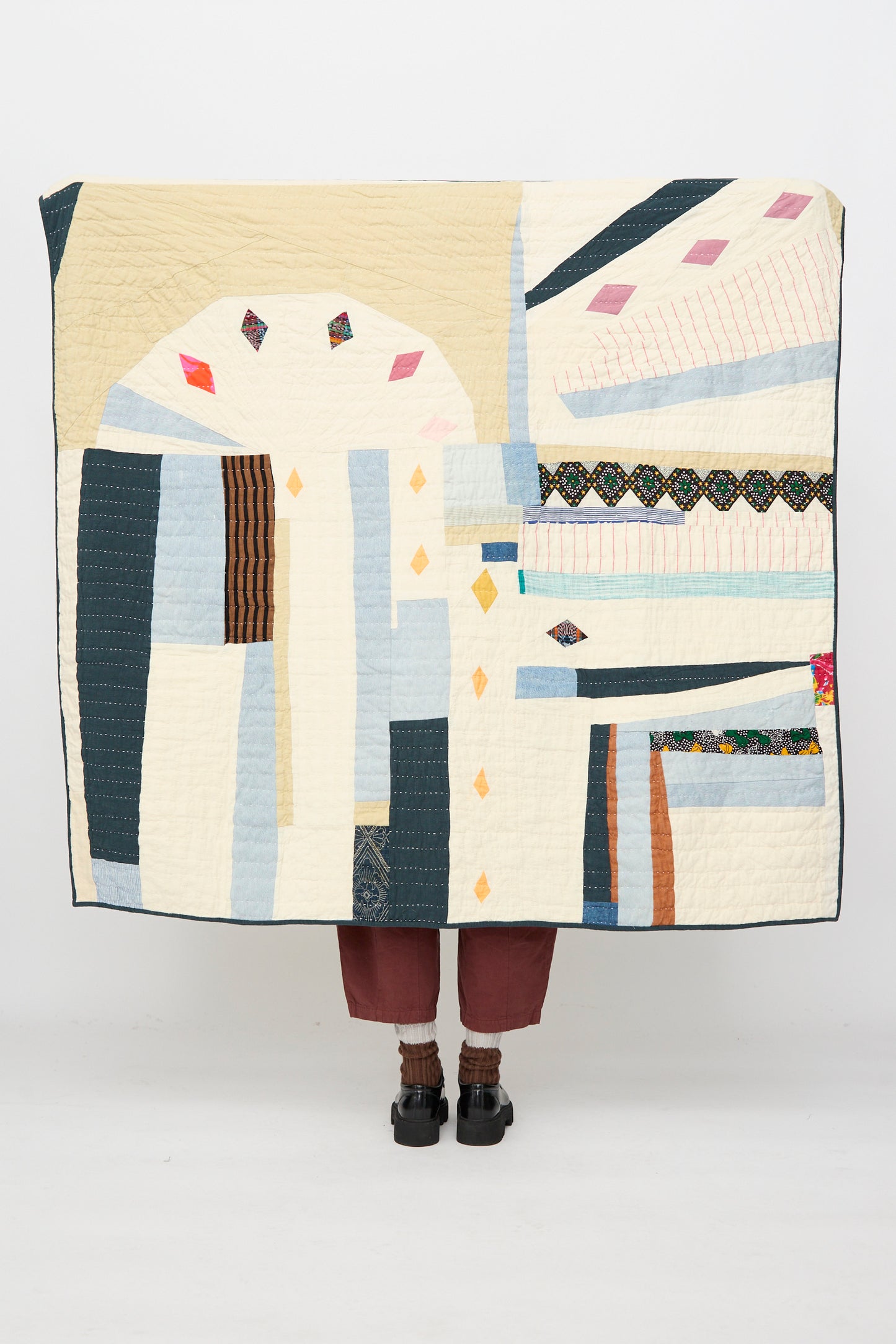 A person holds the Counterpane Large Patchwork Quilt in Cream Multi, handmade from found cloth, with only their lower legs and feet visible.