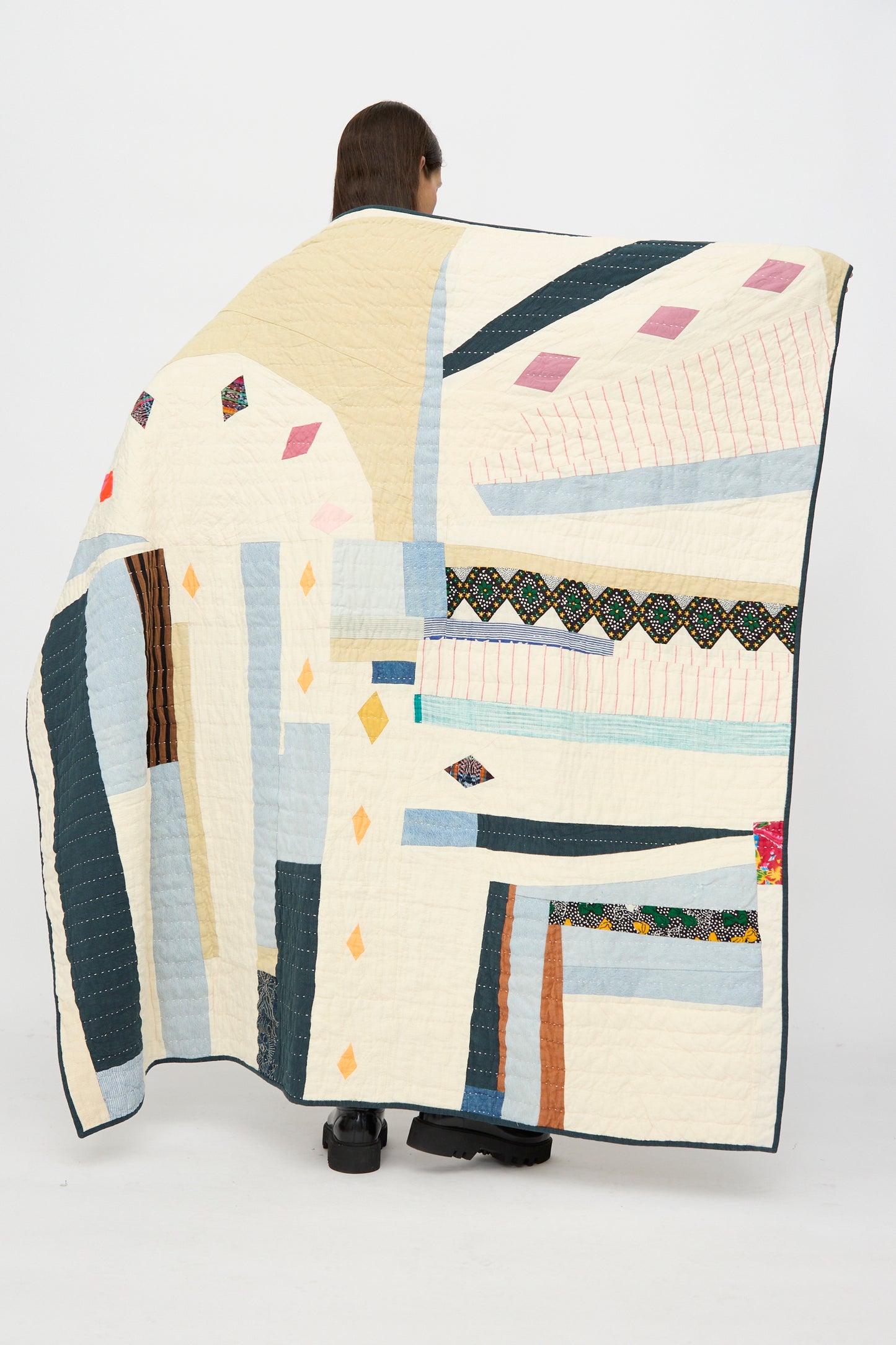 A person stands holding the Counterpane Large Patchwork Quilt in Cream Multi, featuring geometric patterns and various fabric textures, partially draped over their body.