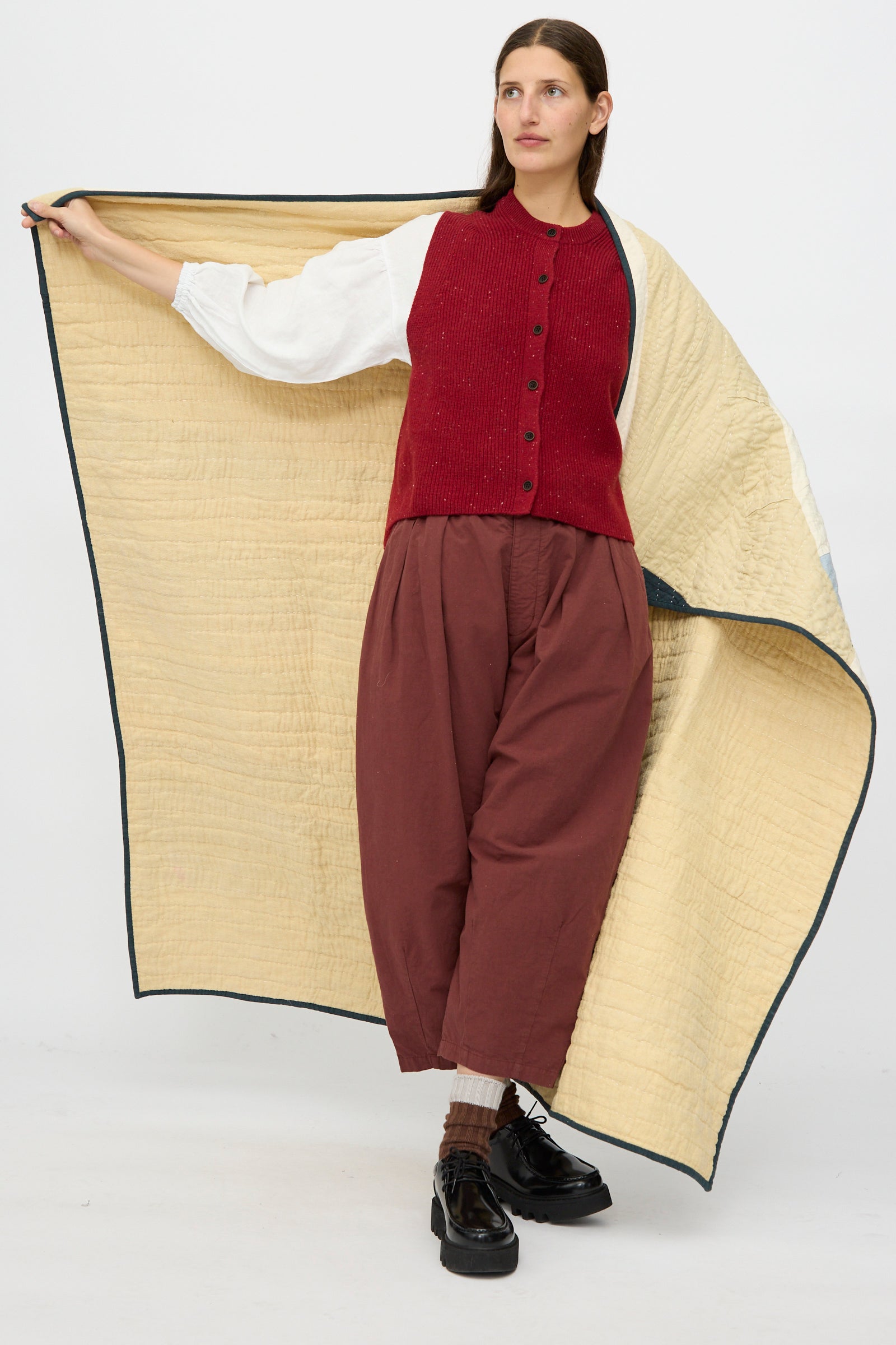 A person in wide maroon pants, a red buttoned vest, and black shoes holds out the Counterpane Large Patchwork Quilt in Cream Multi against a plain white background.