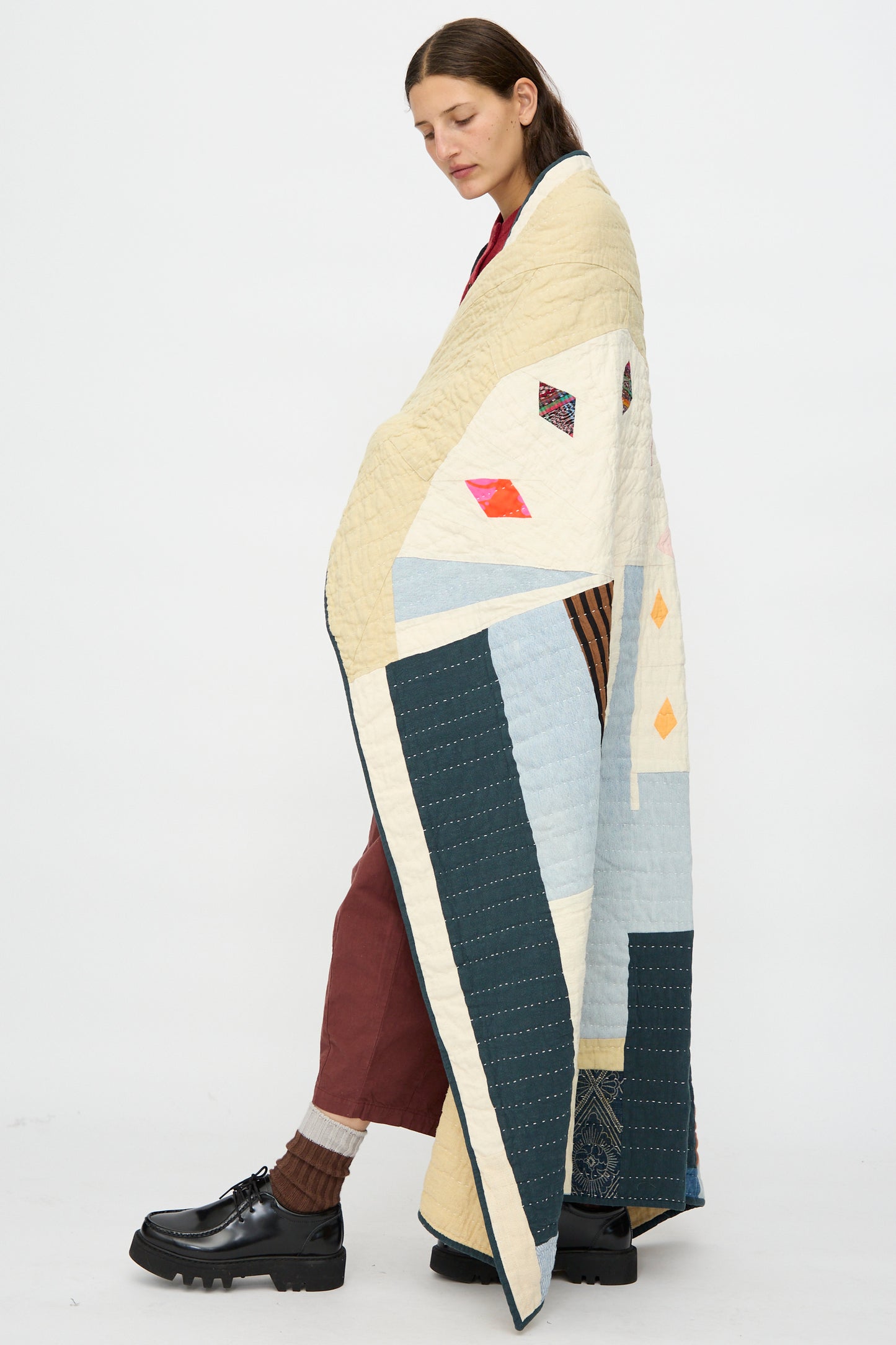 A person stands wrapped in the Counterpane Large Patchwork Quilt in Cream Multi, wearing maroon pants, brown socks, black shoes, and a red shirt against a plain white background.