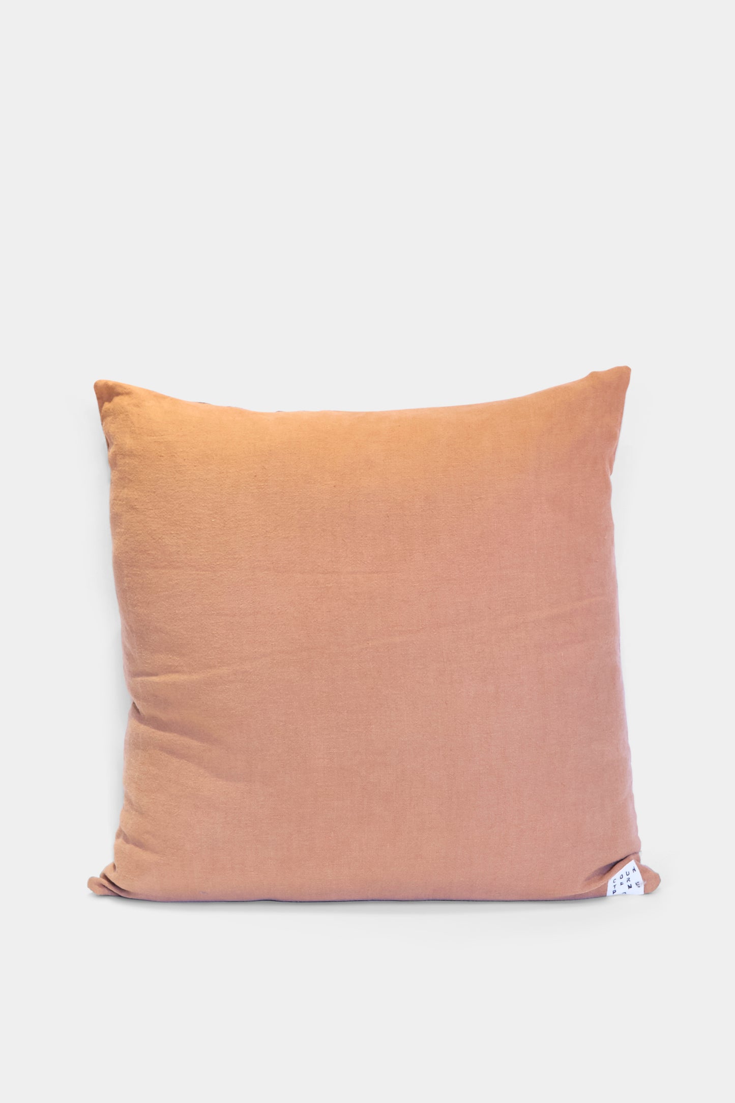 The Counterpane Patchwork Pillow in Denim and Terracotta Multi I is a square, quilted throw pillow in light orange tones with a plain cover and small white tag at the bottom right, beautifully crafted in California, shown on a white background.