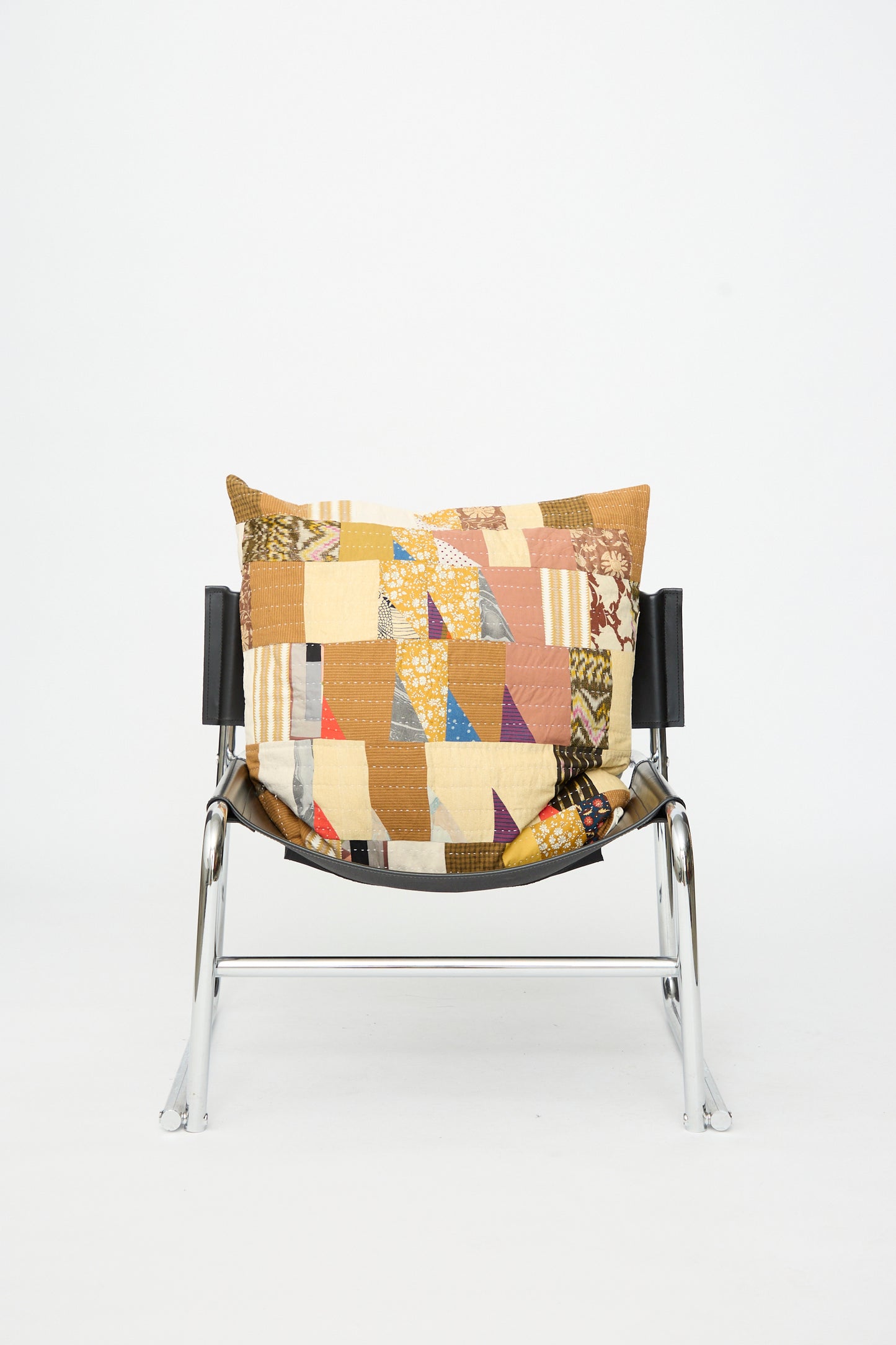 A metal chair with a black seat holds the Counterpane Patchwork Pillow in Earth Multi I, set against a plain white background.