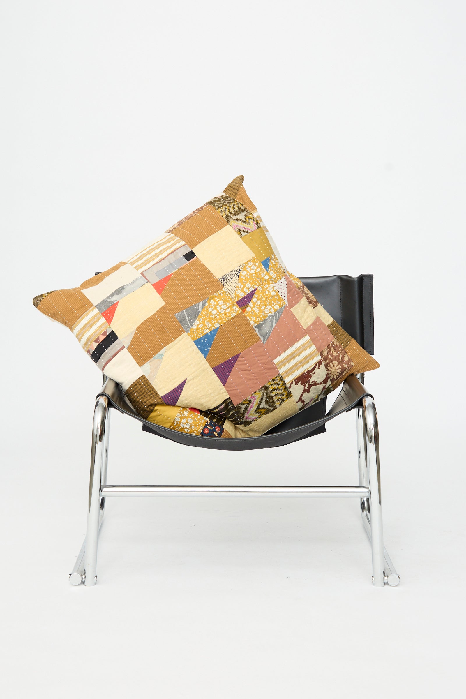 A Counterpane Patchwork Pillow in Earth Multi I, crafted from antique cloth and made in California, rests on a modern black and chrome chair against a plain white background.