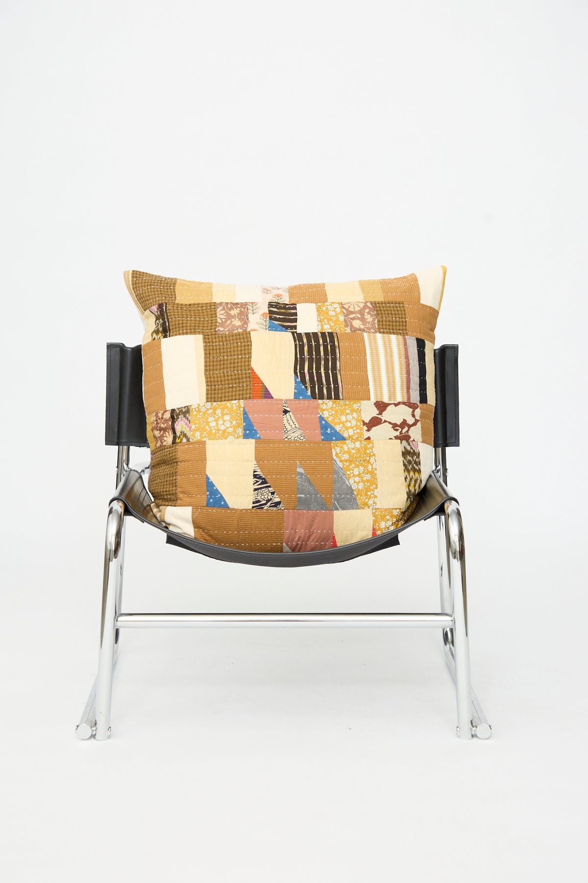 A metal-framed chair with a black seat holds the Counterpane Patchwork Pillow in Yellow Gold Multi II, featuring a large square design covered in brown, yellow, and patterned patchwork made from antique found cloth.