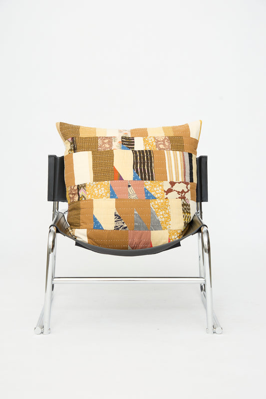 A metal-framed chair with a black seat holds the Counterpane Patchwork Pillow in Yellow Gold Multi II, featuring a large square design covered in brown, yellow, and patterned patchwork made from antique found cloth.
