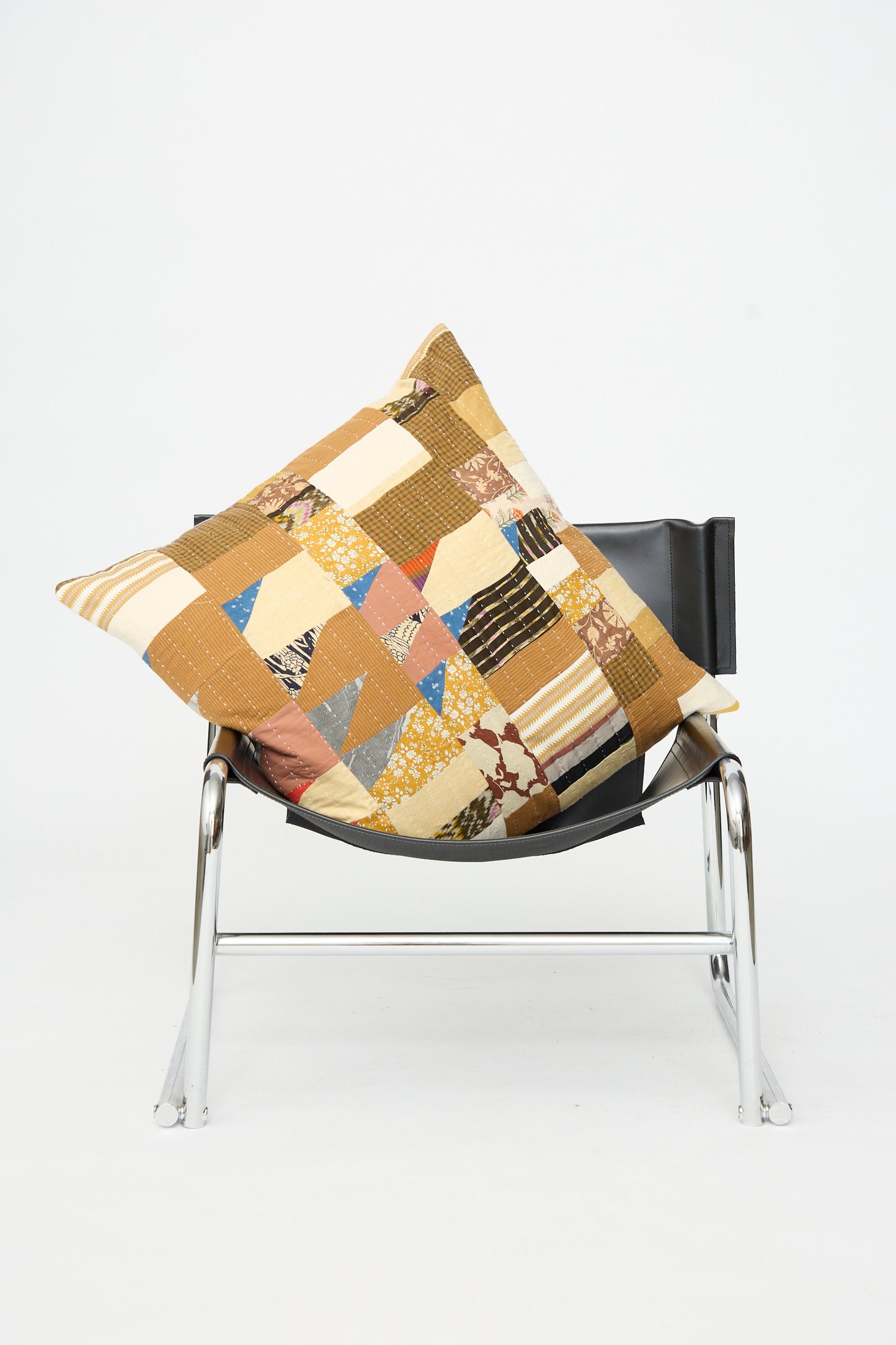 A black chair with a silver metal frame displays the Counterpane Patchwork Pillow in Yellow Gold Multi II—crafted from antique found cloth—resting on its seat, against a plain white background.