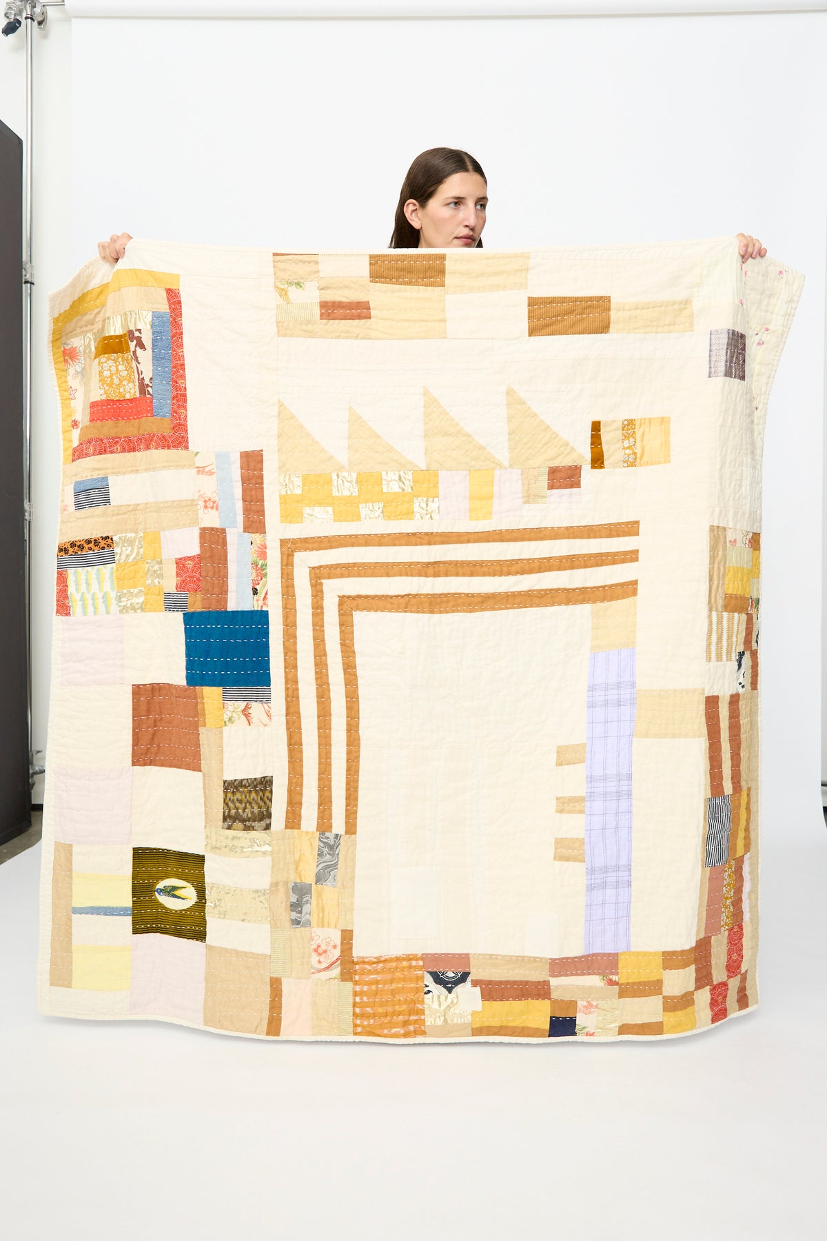 A person holding up the Counterpane Patchwork Quilt in Yellow Gold Multi, featuring handmade geometric patterns in beige, yellow, gold, and pastel hues against a plain studio background.