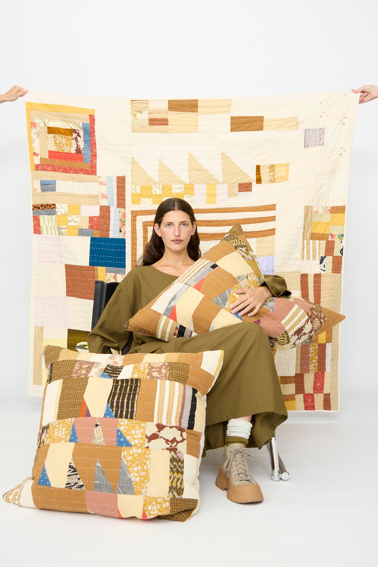 A person in an olive outfit sits holding two patchwork pillows, with the Counterpane Patchwork Quilt in Yellow Gold Multi displayed behind them against a plain white background.