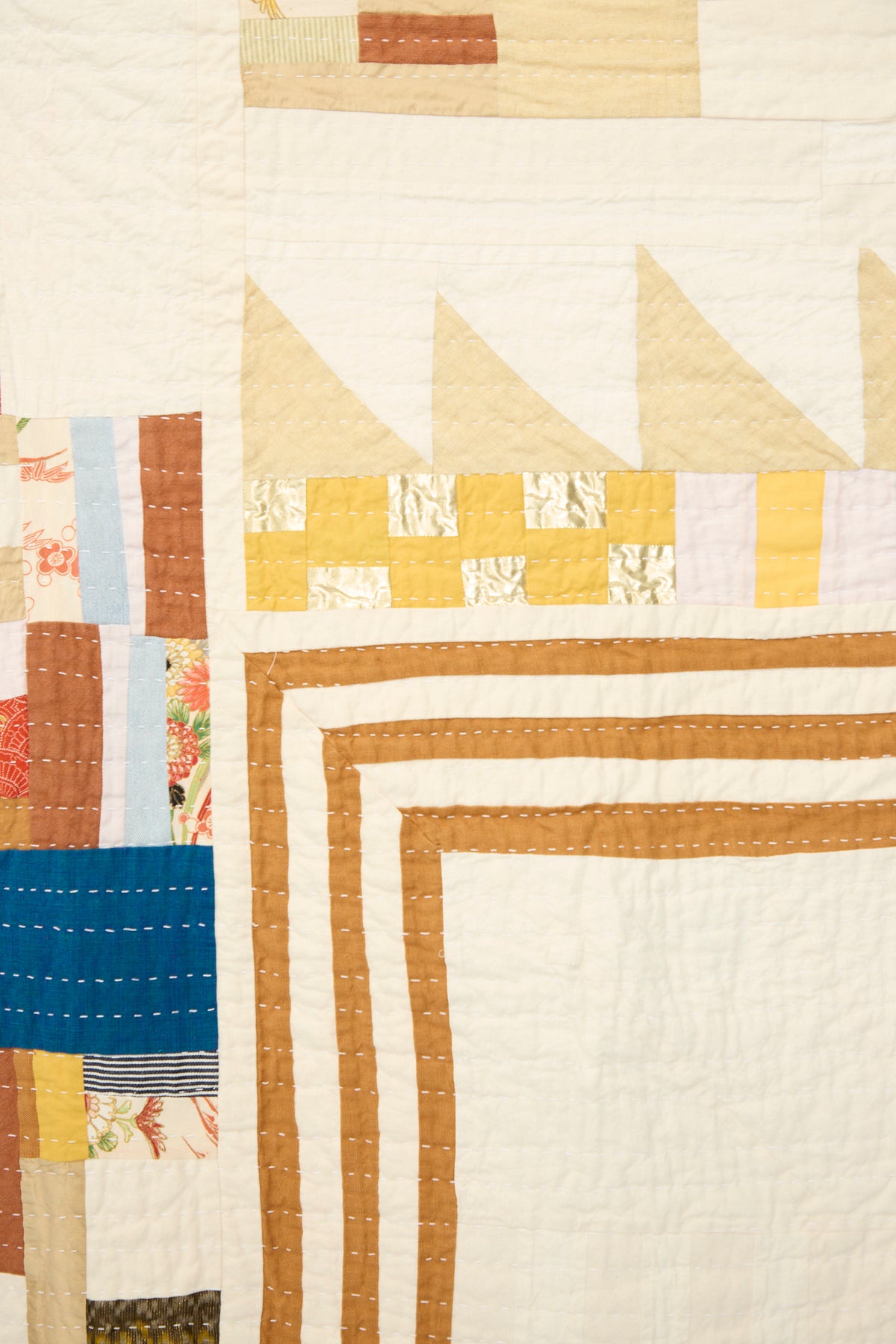 The Counterpane Patchwork Quilt in Yellow Gold Multi showcases geometric shapes—triangles, squares, and rectangles—in warm shades of brown, yellow, blue, and cream. Visible hand stitching highlights the expert craftsmanship of this California-made quilt.
