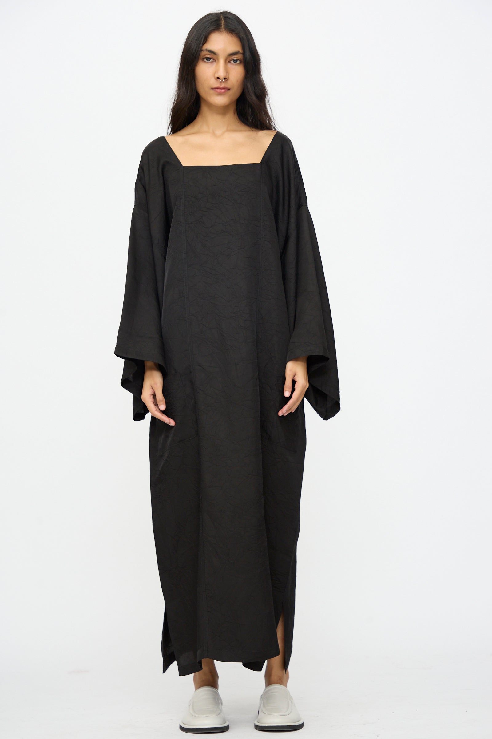 A woman stands facing forward in the Cristaseya Square Neck Caftan Dress in Black—a loose, long dress with wide sleeves—paired with white slip-on shoes against a plain white background.