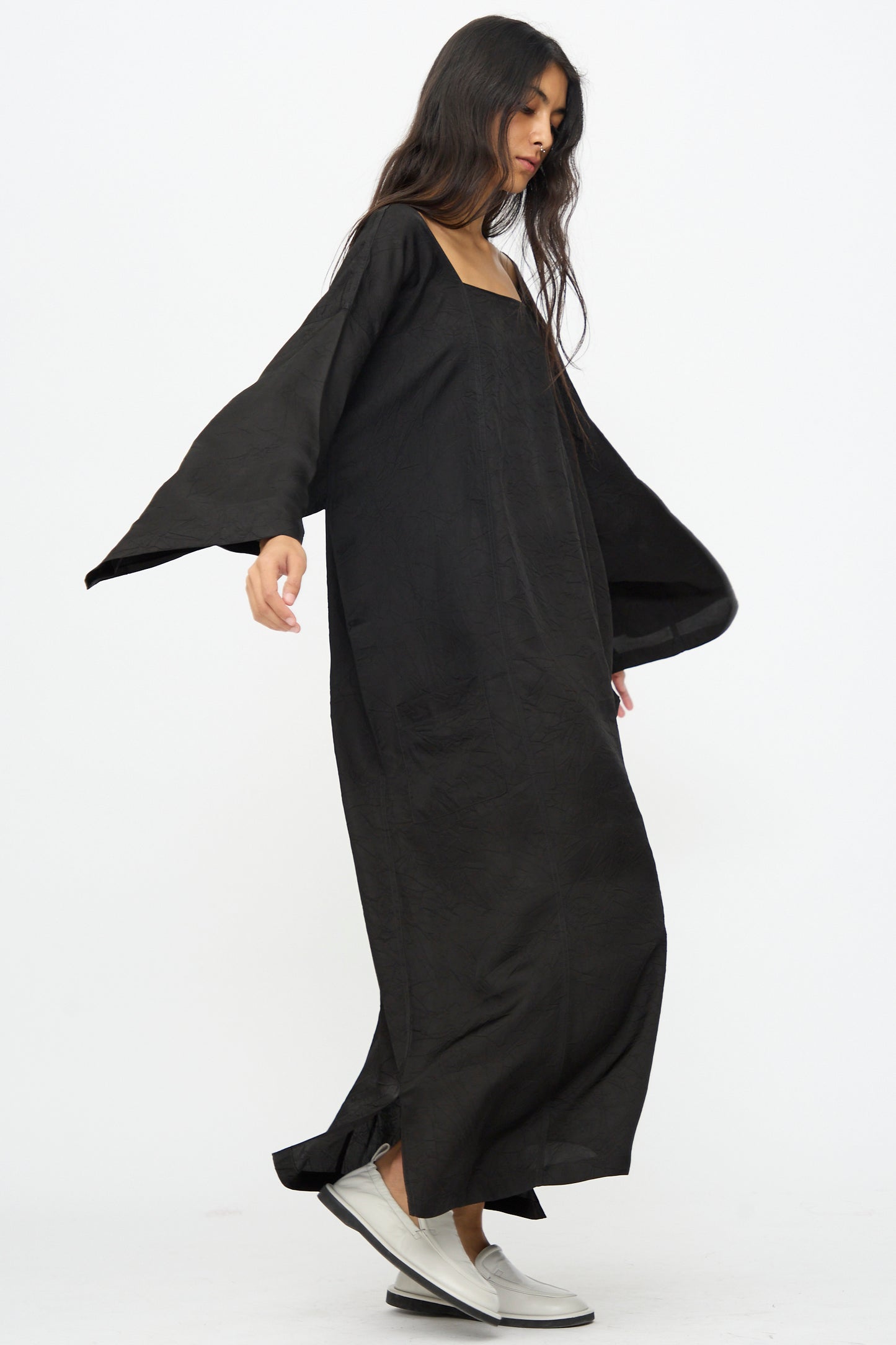 A woman models the Cristaseya Square Neck Caftan Dress in black, a loose-fitting Italian cupro maxi dress with wide sleeves, styled with silver slip-on shoes against a plain white background.