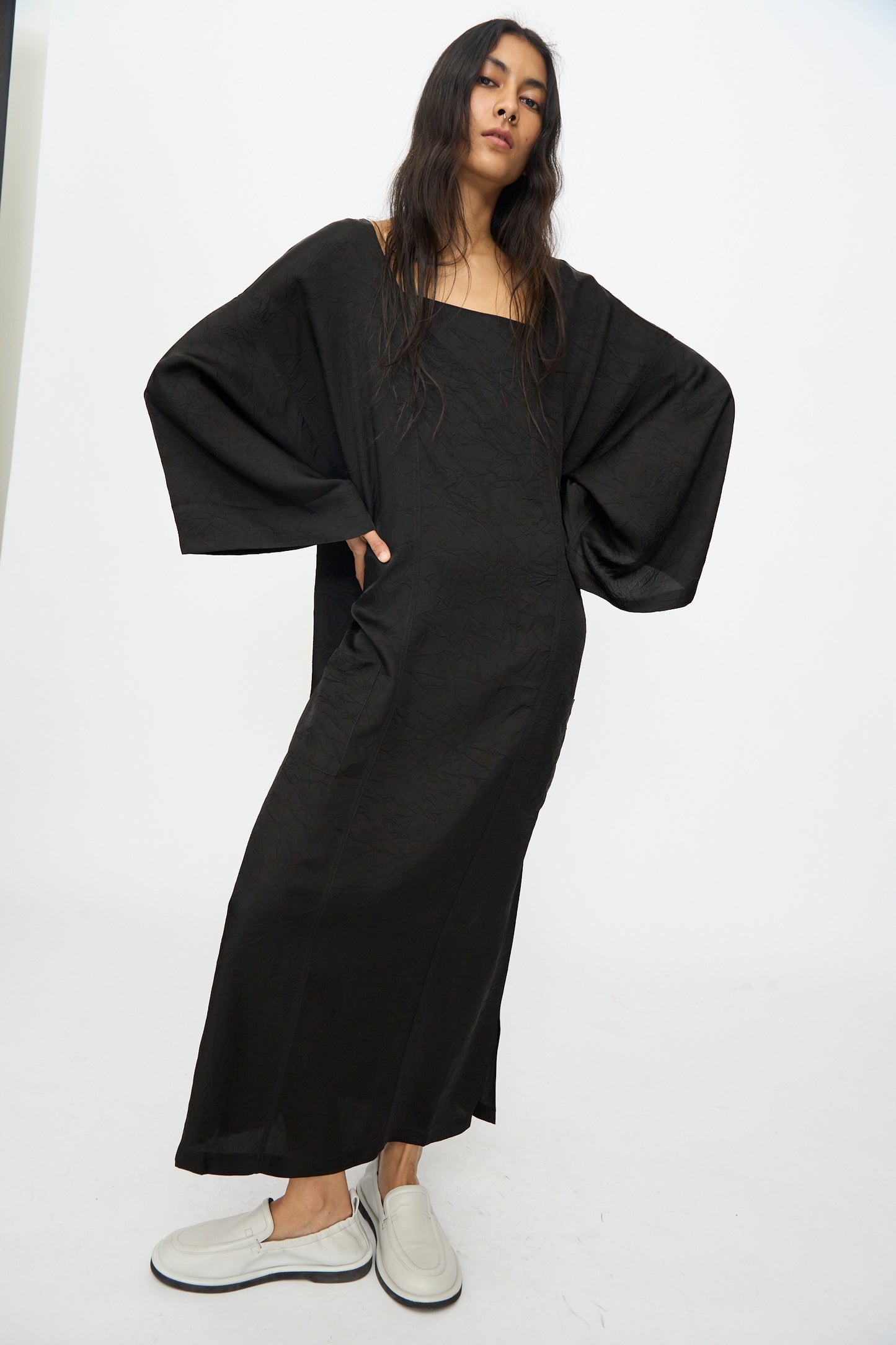 A person stands against a plain background, wearing the Cristaseya Square Neck Caftan Dress in Black with wide sleeves, paired with white slip-on shoes.