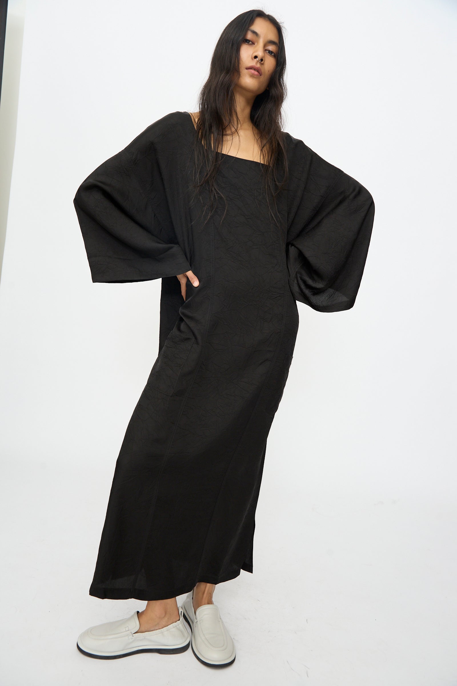 A person stands against a plain background, wearing the Cristaseya Square Neck Caftan Dress in Black with wide sleeves, paired with white slip-on shoes.