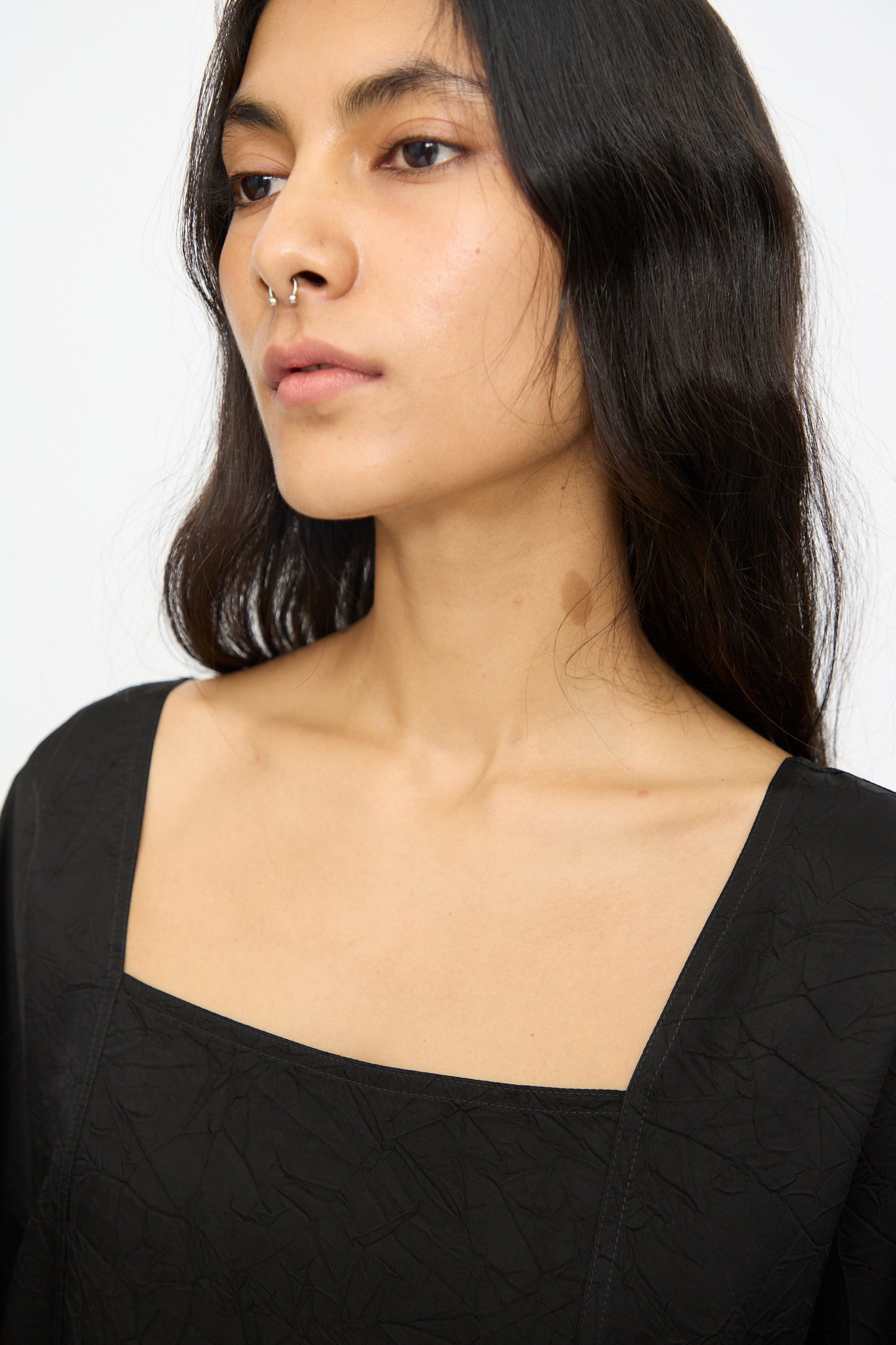 A person with long dark hair, wearing the Cristaseya Square Neck Caftan Dress in Black and a nose ring, gazes slightly to the side against a plain background.