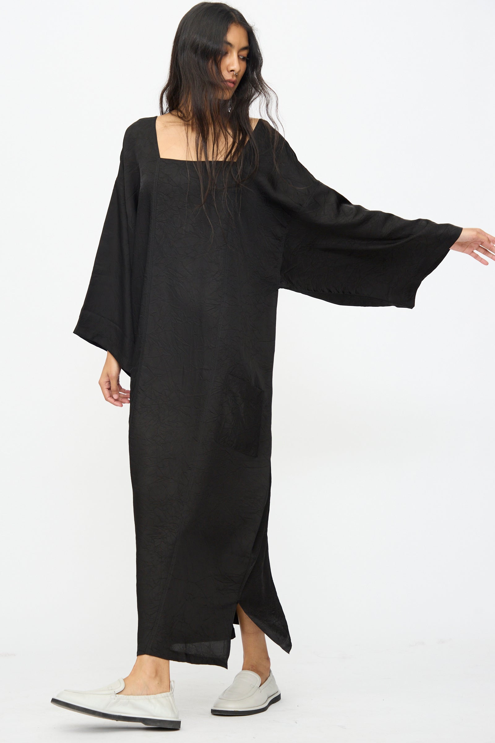 A person wearing the Cristaseya Square Neck Caftan Dress in Black with wide sleeves and white slip-on shoes stands against a plain white background.
