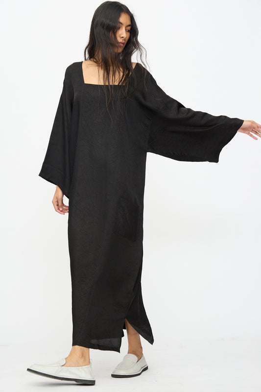A person wearing the Cristaseya Square Neck Caftan Dress in Black with wide sleeves and white slip-on shoes stands against a plain white background.